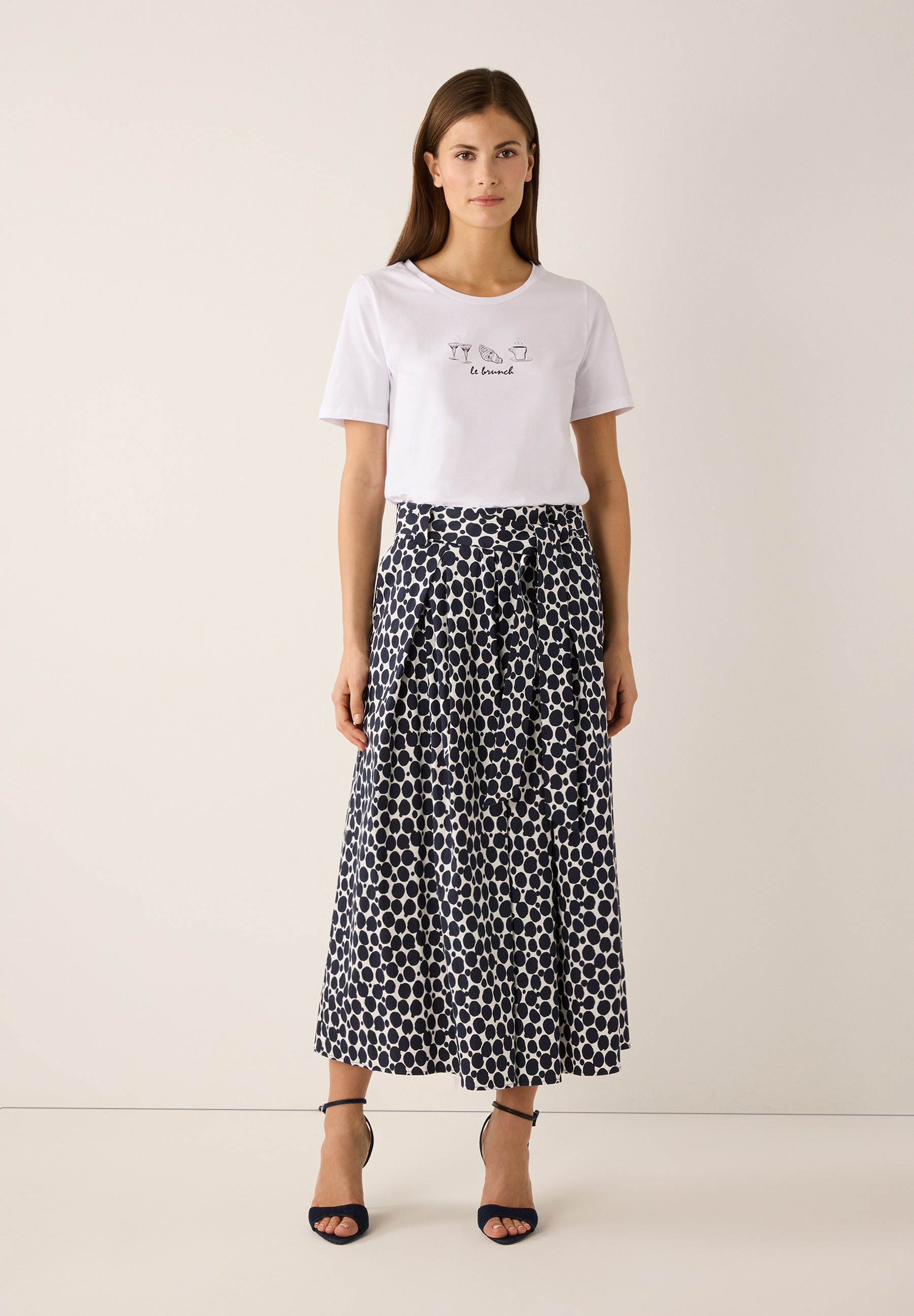 Eine Frau im MORE & MORE T-Shirt mit "Le Brunch"-Aufdruck und einem Rock mit Punktmuster. Eine Frau im MORE & MORE T-Shirt mit "Le Brunch"-Aufdruck und einem Rock mit Punktmuster.