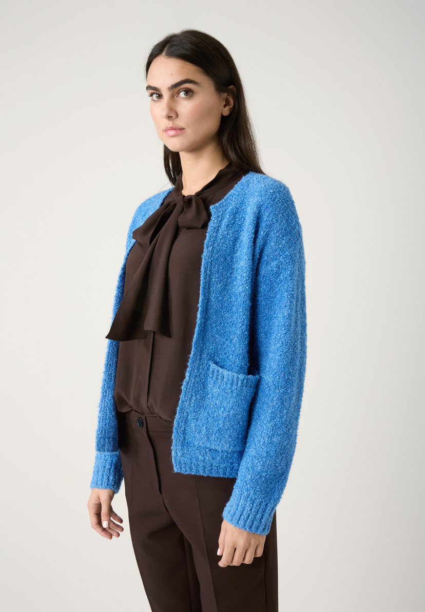 Ein MORE & MORE Strick-Oberteil aus Bouclé mit angenehmem Tragekomfort und weichem Griff, kombiniert mit einer braunen Bluse und Hose. Ein MORE & MORE Strick-Oberteil aus Bouclé mit angenehmem Tragekomfort und weichem Griff, kombiniert mit einer braunen Bluse und Hose.