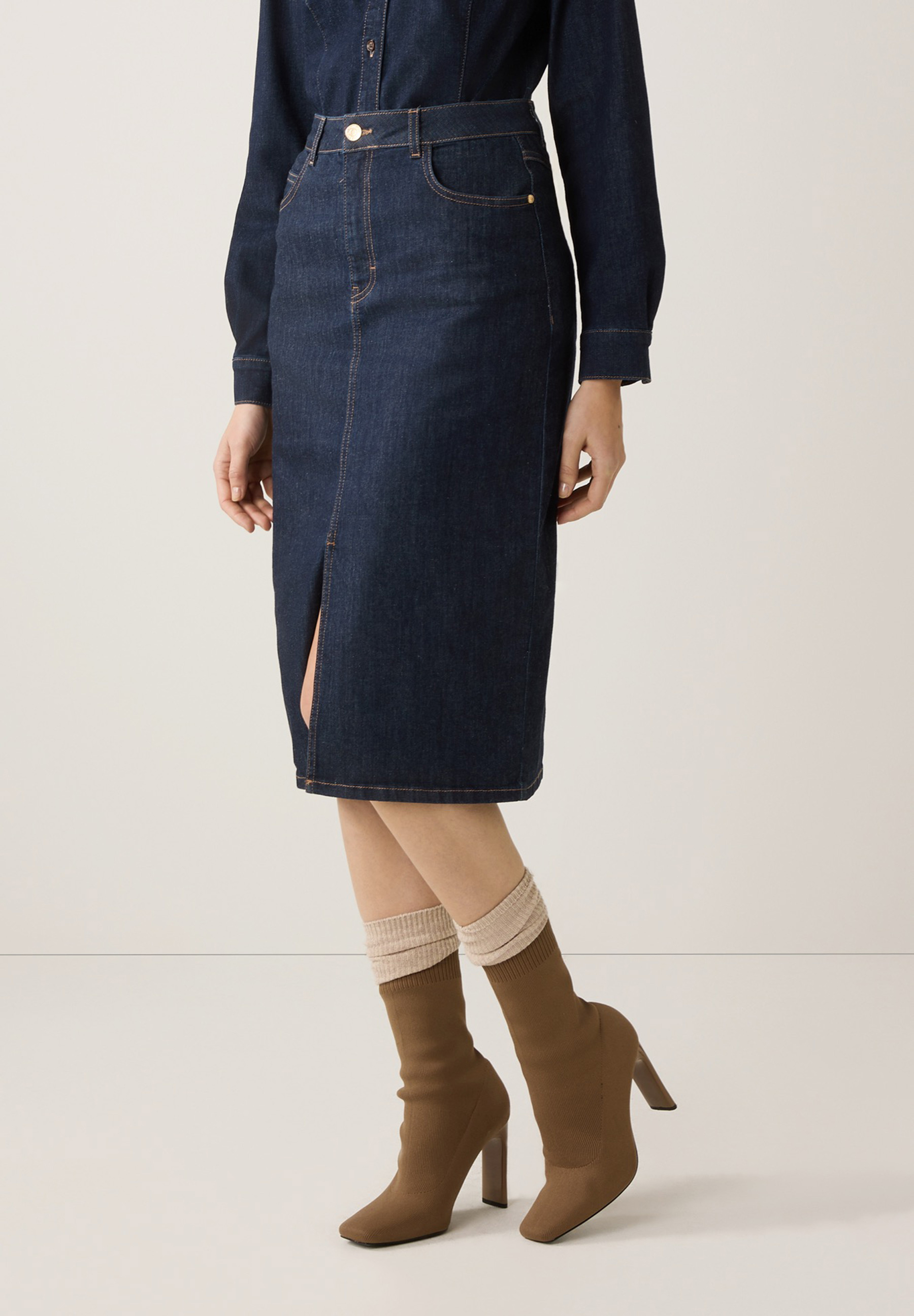 Eine Person trägt einen MORE & MORE Rock aus Denim mit Schlitz. Dazu braune Stiefel mit Absatz.