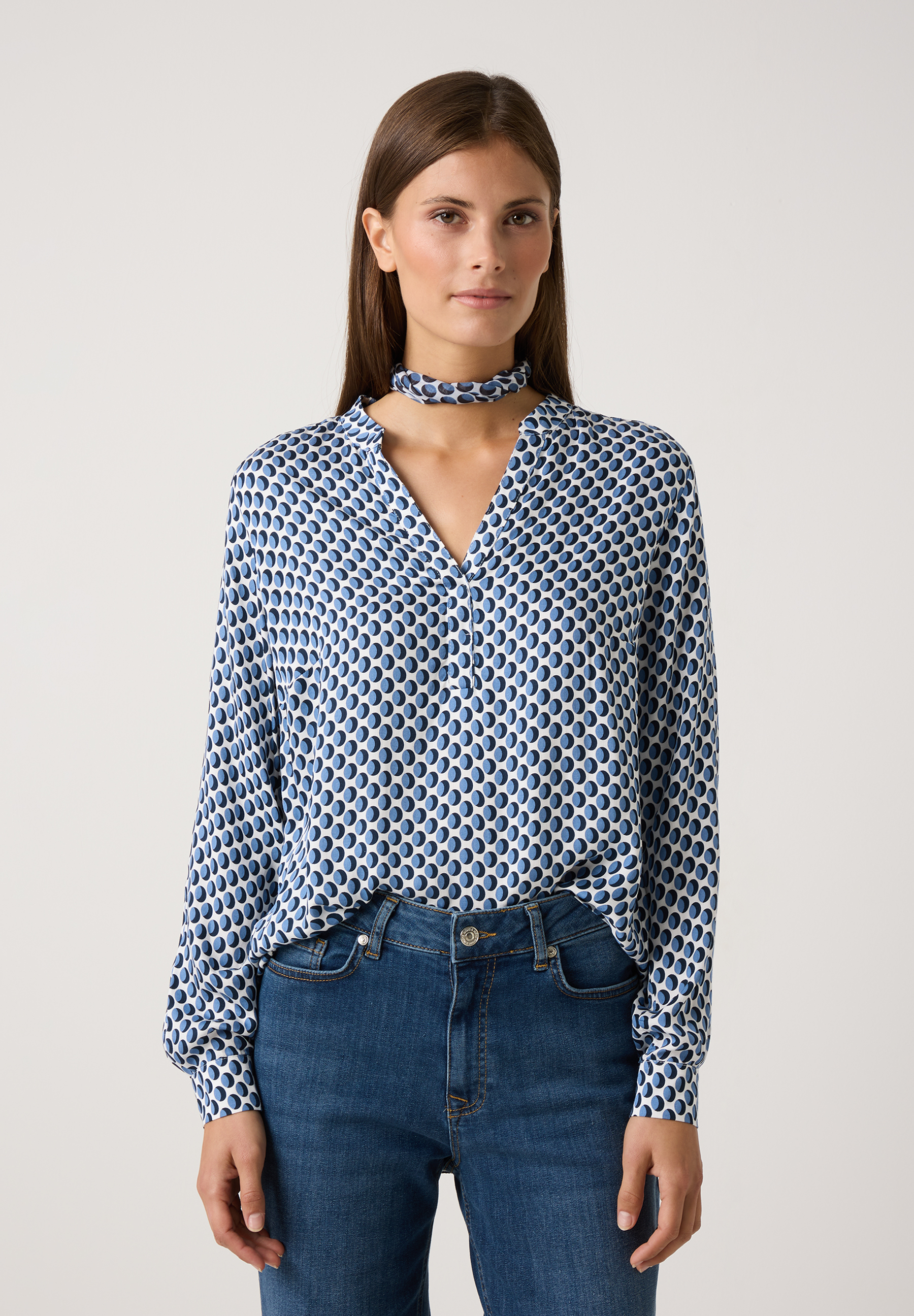 Eine Frau mit einer MORE & MORE Bluse aus Viskose mit blau-weißem Punktmuster und passendem Halsband, kombiniert mit Jeans.