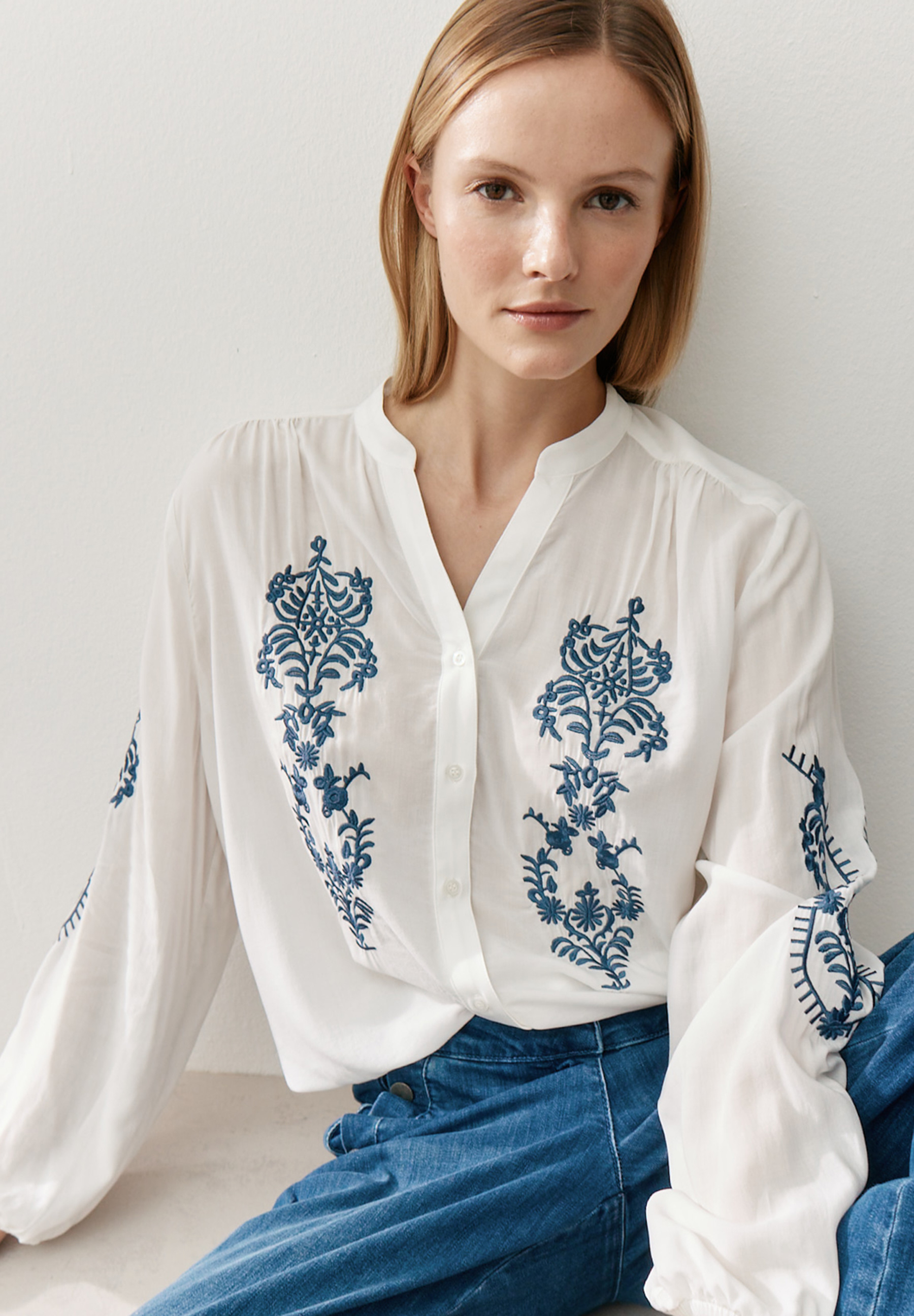 Eine Frau in einer weißen MORE & MORE Bluse mit blauen Stickereien und Jeans.