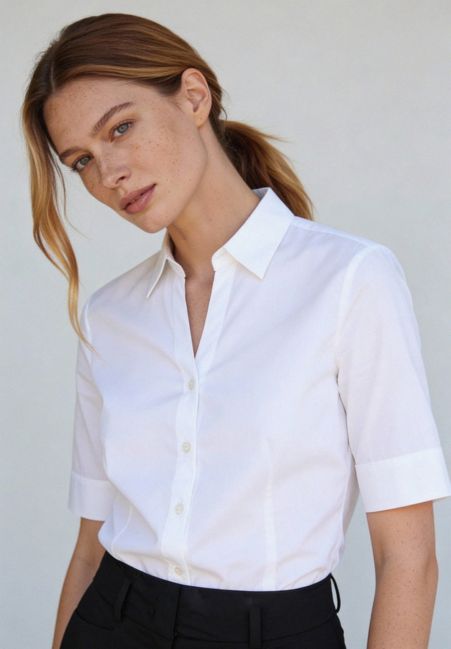 Eine Frau mit Sommersprossen, die eine weiße MORE & MORE Bluse aus Baumwollmischung trägt, blickt in die Kamera.