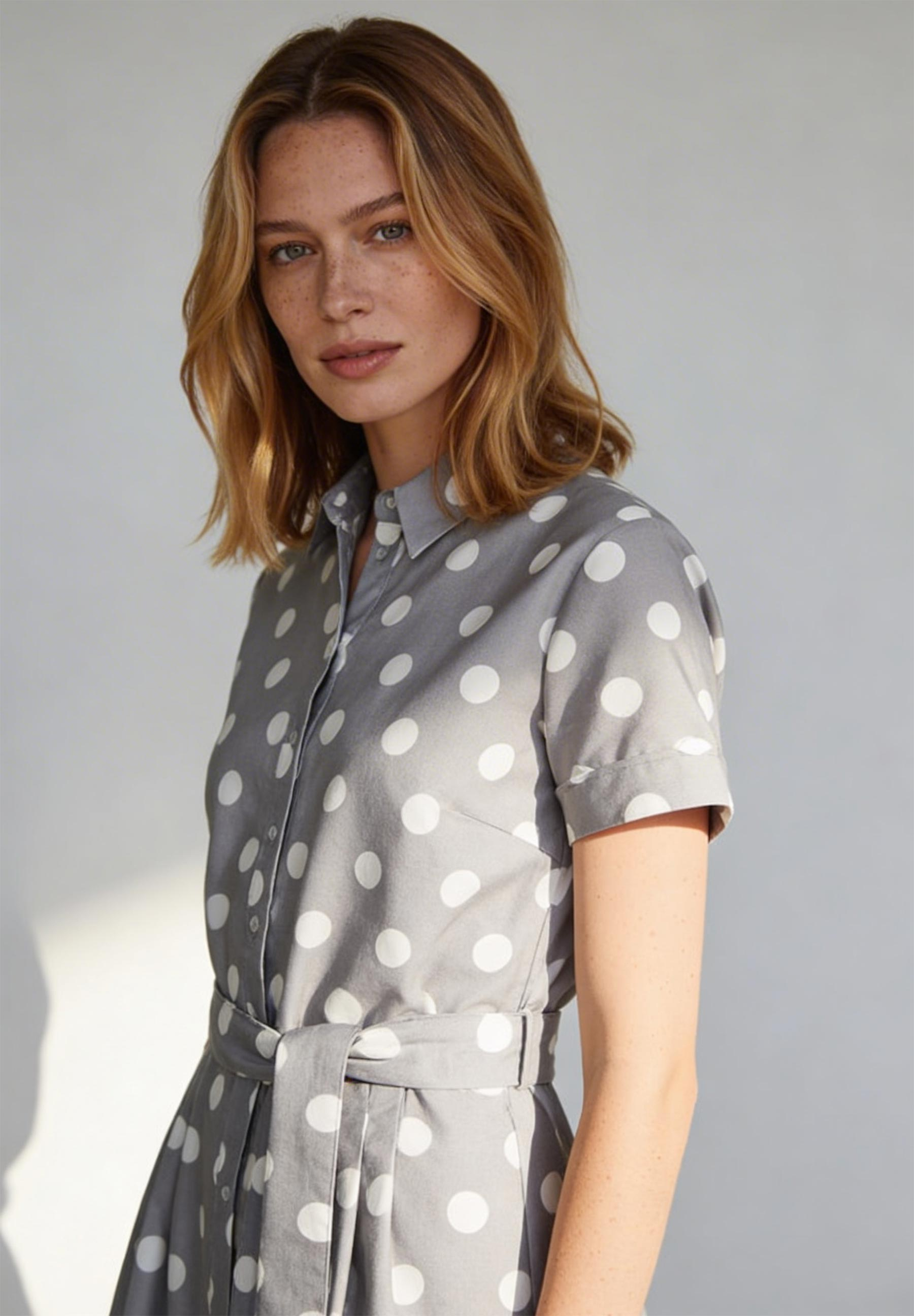 Eine Frau, die ein graues MORE & MORE Oberteil mit weißen Punkten und Gürtel trägt.