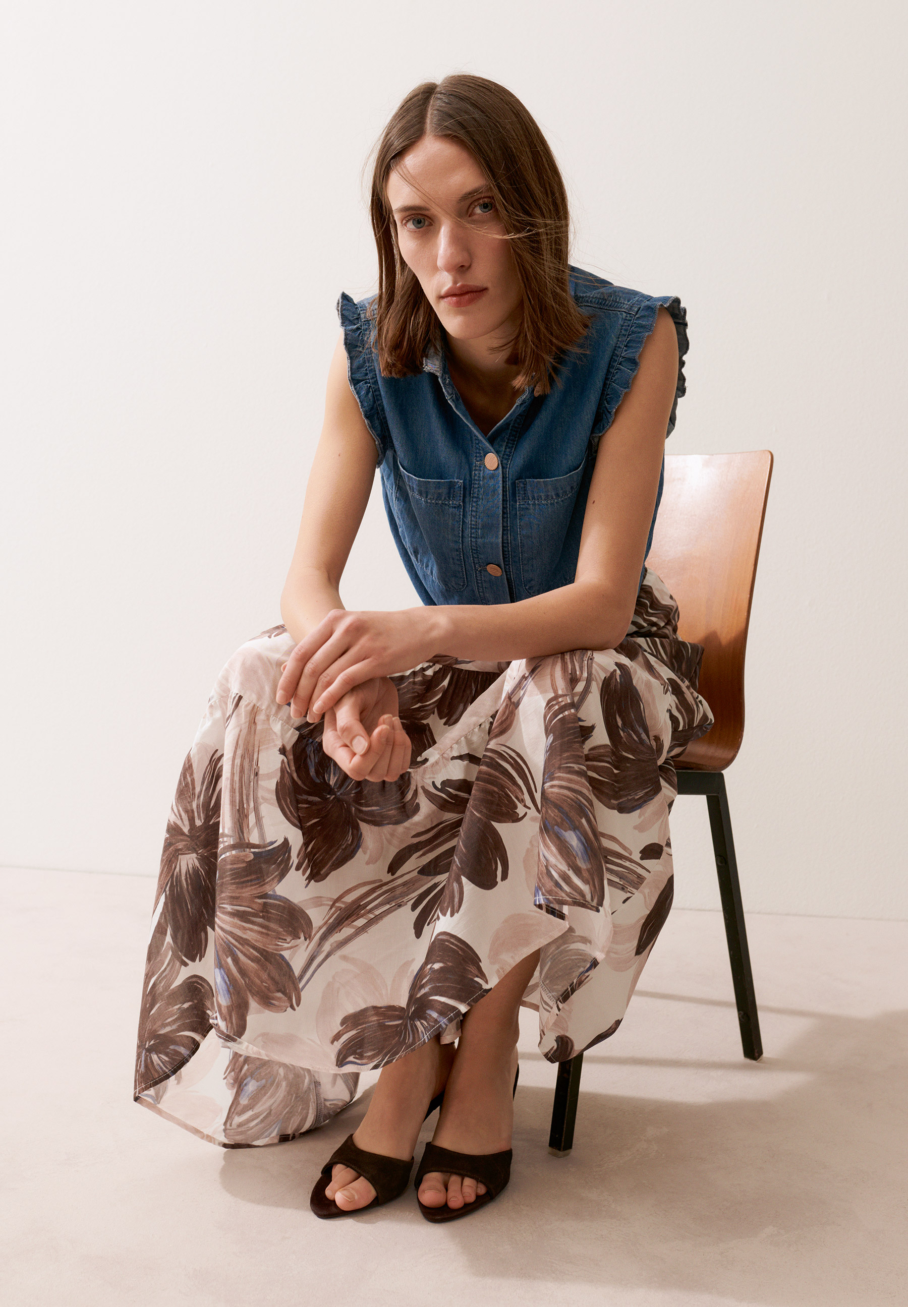 Eine Frau in einer MORE & MORE Bluse mit floralem Rock sitzt auf einem Holzstuhl.