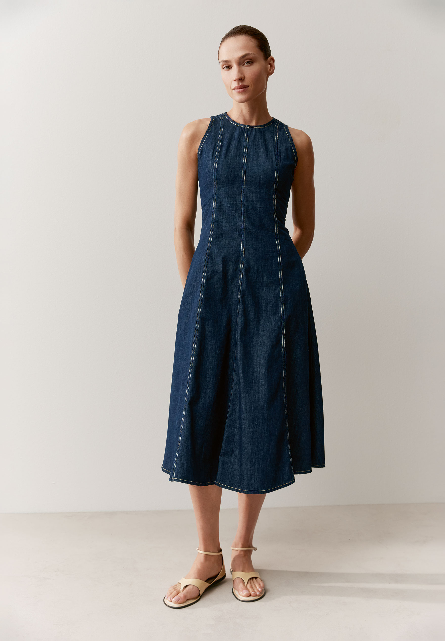Eine Frau in einem ärmellosen Jeanskleid von MORE & MORE mit A-Linien-Schnitt und weißen Ziernähten.