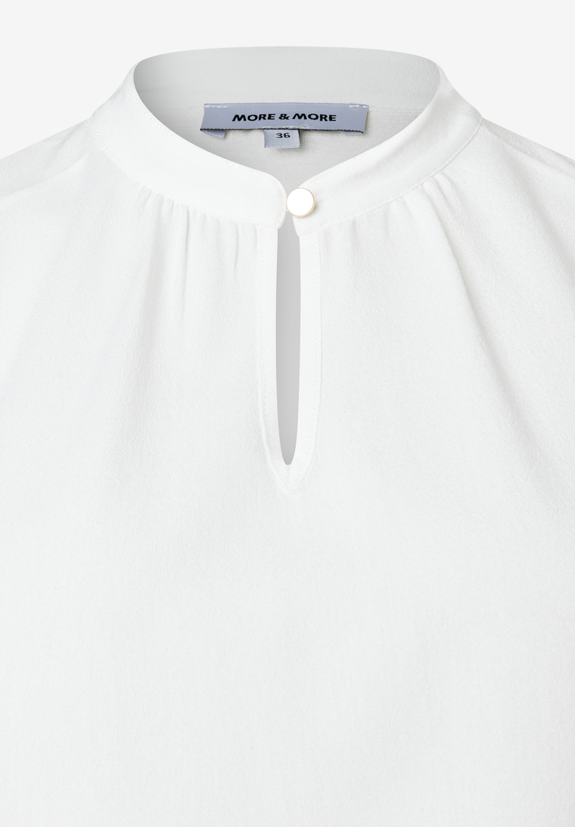 Nahaufnahme einer weißen MORE & MORE Bluse mit Stehkragen und Knopfdetail. Nahaufnahme einer weißen MORE & MORE Bluse mit Stehkragen und Knopfdetail.