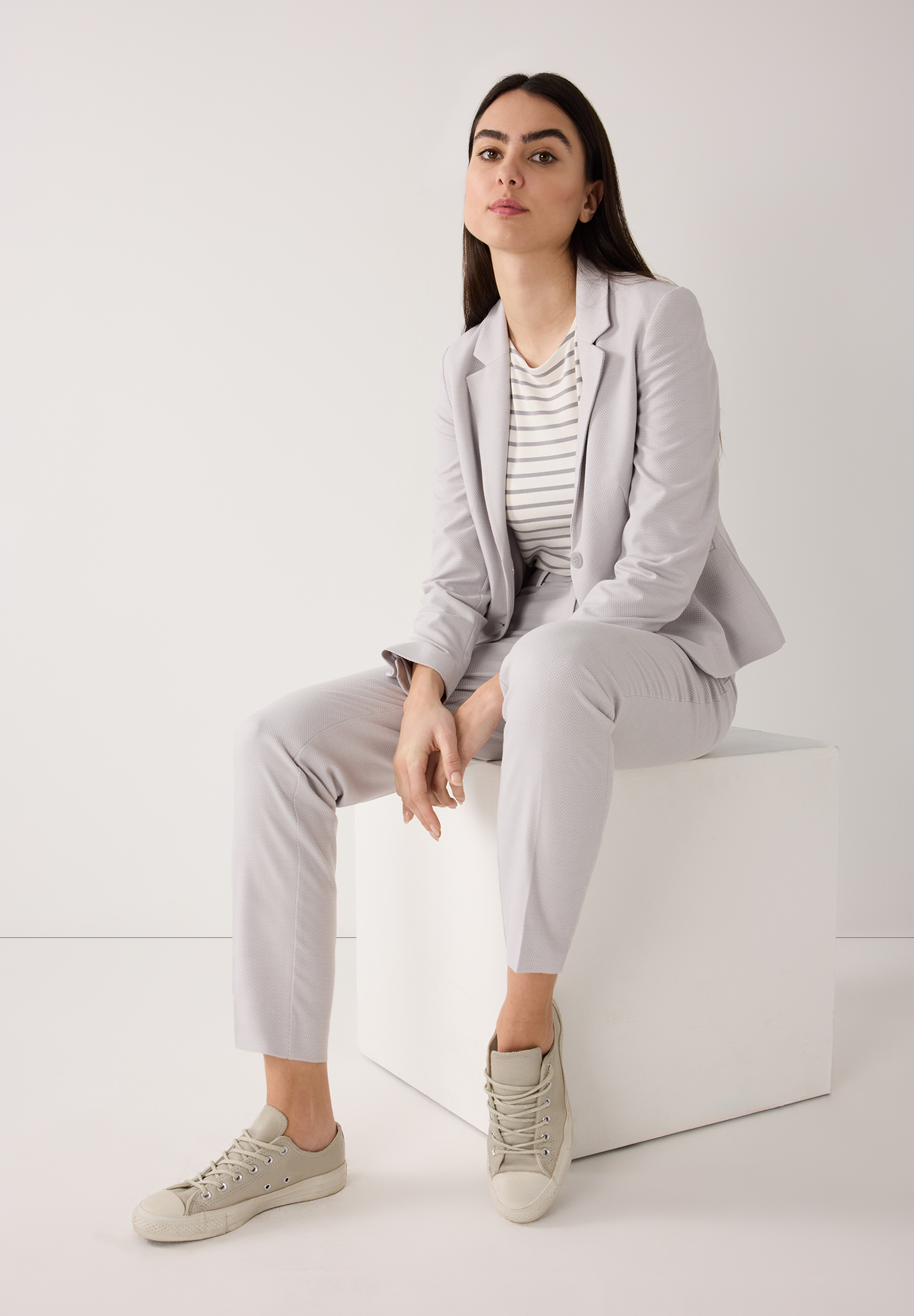 Eine Frau sitzt in einem MORE & MORE Blazeranzug aus Piqué-Stretch mit Sneakern vor einem weißen Hintergrund. Eine Frau sitzt in einem MORE & MORE Blazeranzug aus Piqué-Stretch mit Sneakern vor einem weißen Hintergrund.