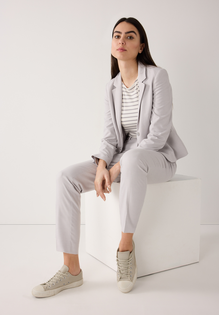 Eine Frau sitzt in einem MORE & MORE Blazeranzug aus Piqué-Stretch mit Sneakern vor einem weißen Hintergrund. Eine Frau sitzt in einem MORE & MORE Blazeranzug aus Piqué-Stretch mit Sneakern vor einem weißen Hintergrund.
