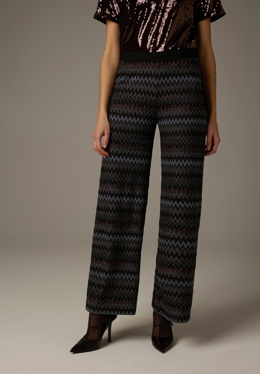 Eine Person mit MORE & MORE Jersey-Hosen mit Zickzackmuster und High Heels. Eine Person mit MORE & MORE Jersey-Hosen mit Zickzackmuster und High Heels.
