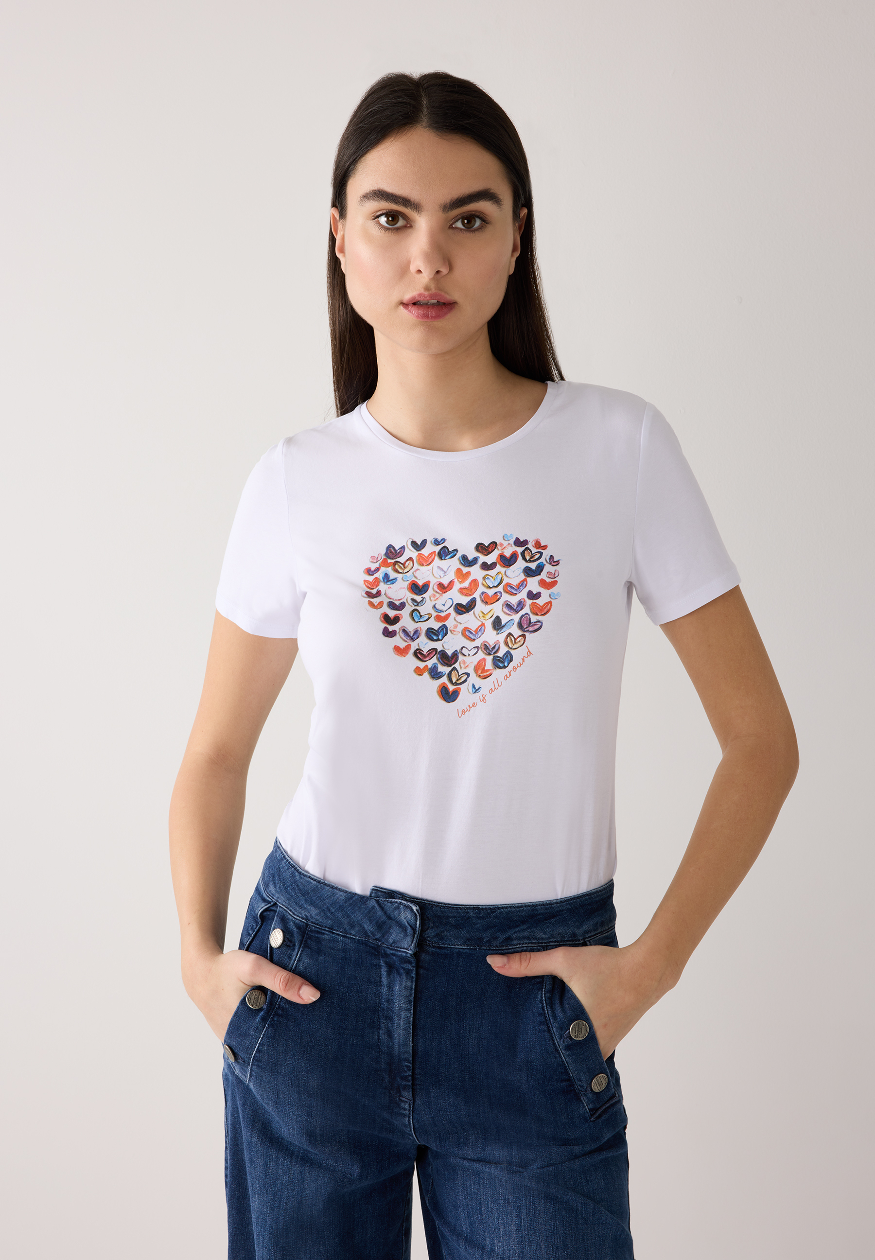 Eine Frau trägt ein weißes MORE & MORE T-Shirt mit Herzmuster und blaue Jeans.