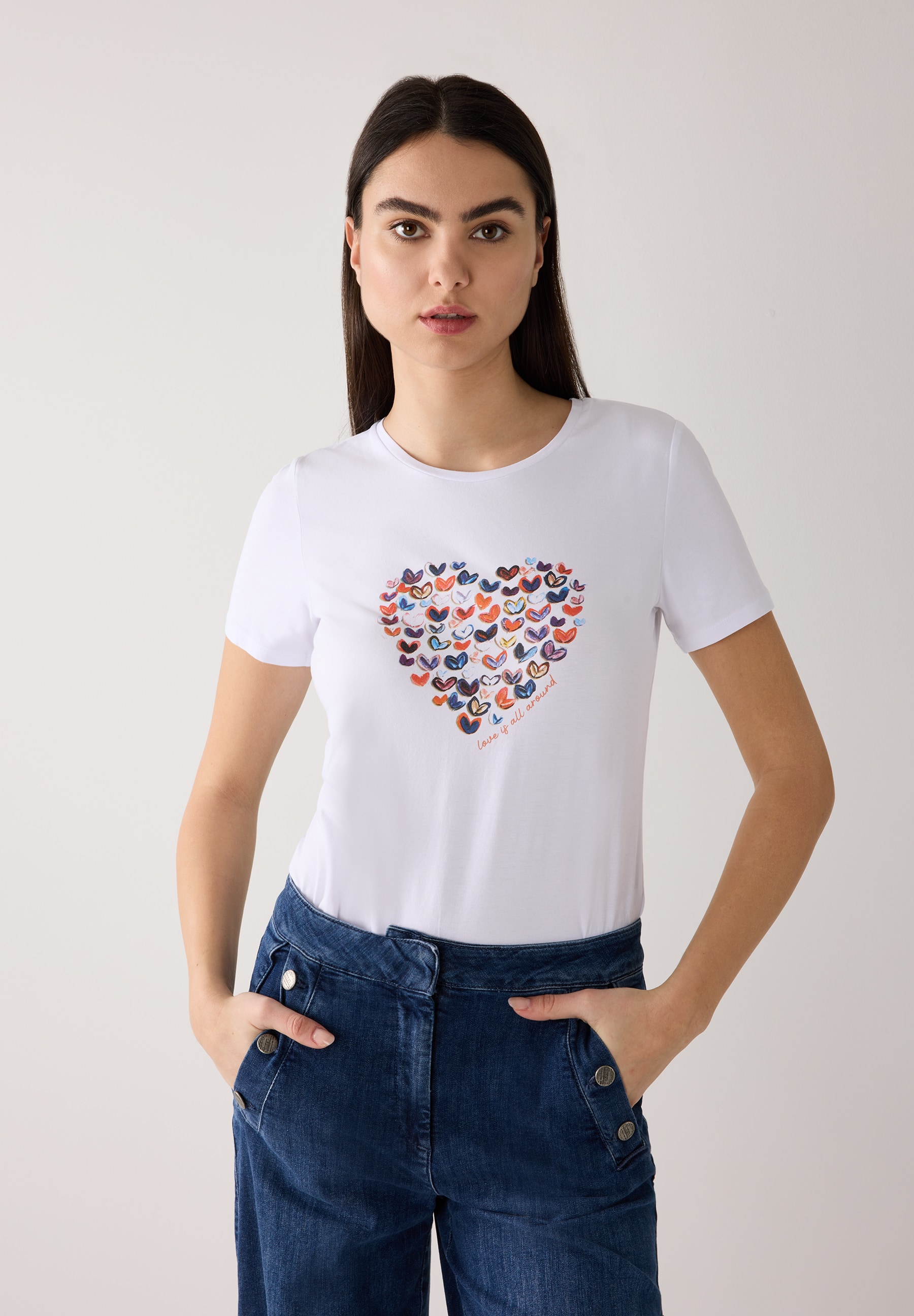 Eine Frau trägt ein weißes MORE & MORE T-Shirt mit Herzmuster und blaue Jeans. Eine Frau trägt ein weißes MORE & MORE T-Shirt mit Herzmuster und blaue Jeans.