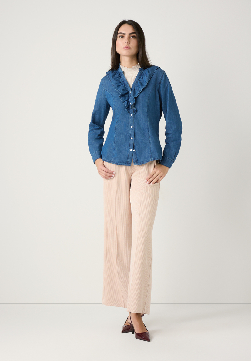 Eine Frau in MORE & MORE Oberteilen, einer Jeansbluse mit Rüschen und beigen Cordhosen. Sie posiert vor einem weißen Hintergrund. Eine Frau in MORE & MORE Oberteilen, einer Jeansbluse mit Rüschen und beigen Cordhosen. Sie posiert vor einem weißen Hintergrund.