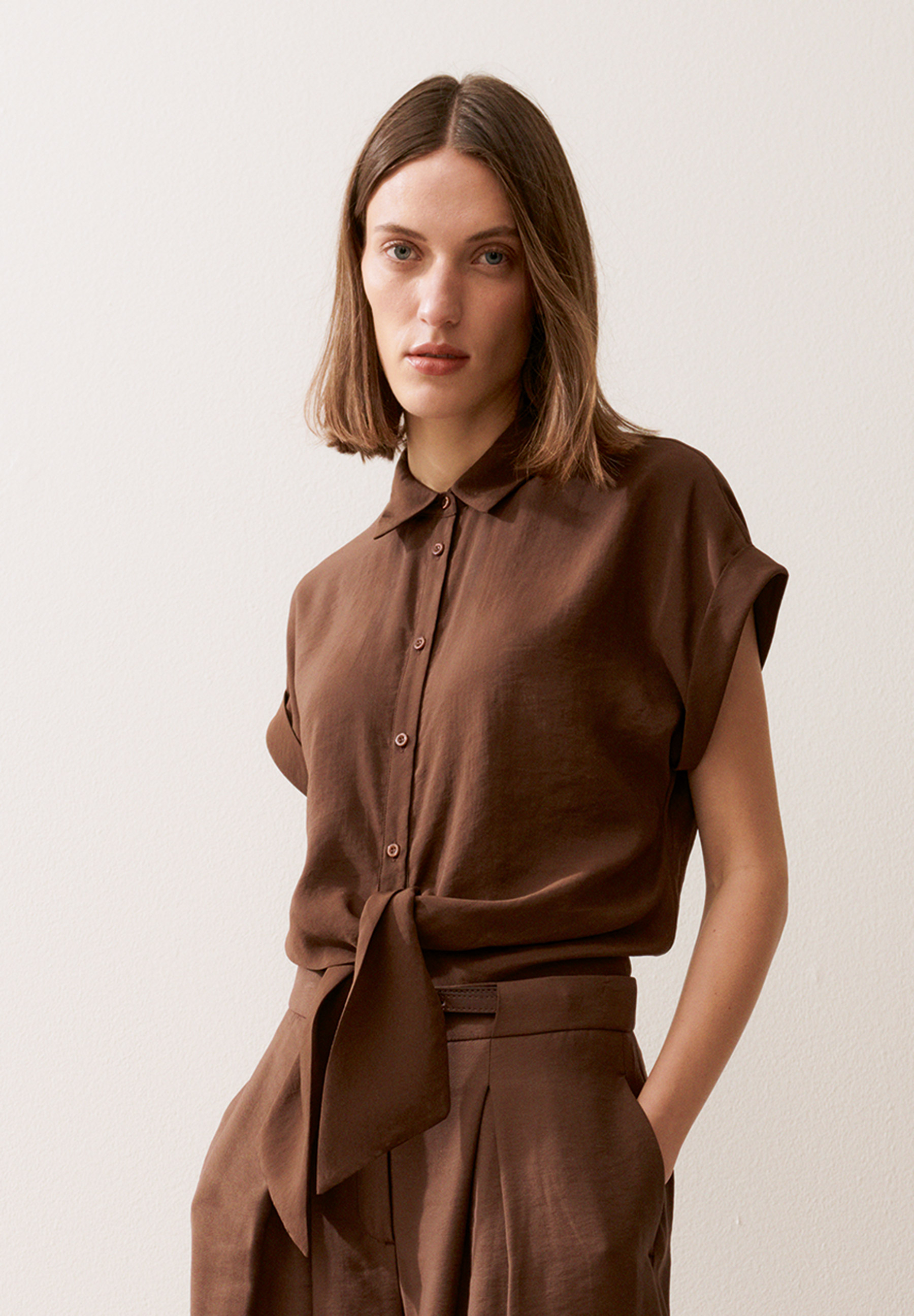 Eine Frau trägt eine MORE & MORE Bluse mit Knopfleiste und Knotendetail in Braun.