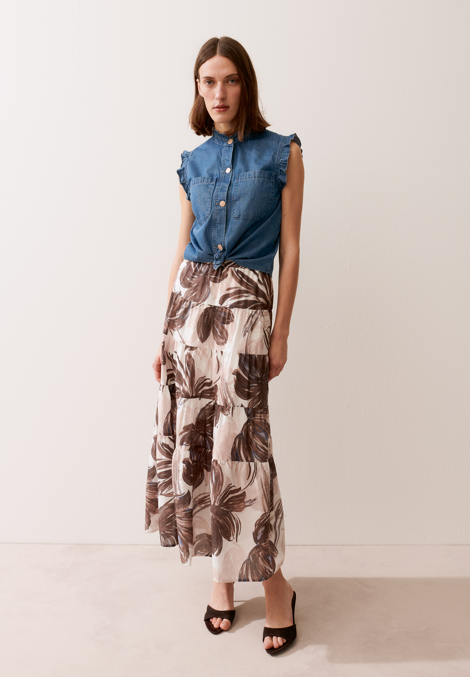 Eine Frau trägt eine MORE & MORE Bluse aus Jeansstoff mit Rüschenärmeln und einen langen, geblümten Rock.