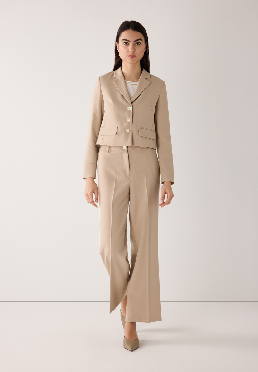 Eine Frau in einem beigen Hosenanzug von MORE & MORE. Die Webware-Hose ist fließend mit hohem Tragekomfort. Eine Frau in einem beigen Hosenanzug von MORE & MORE. Die Webware-Hose ist fließend mit hohem Tragekomfort.