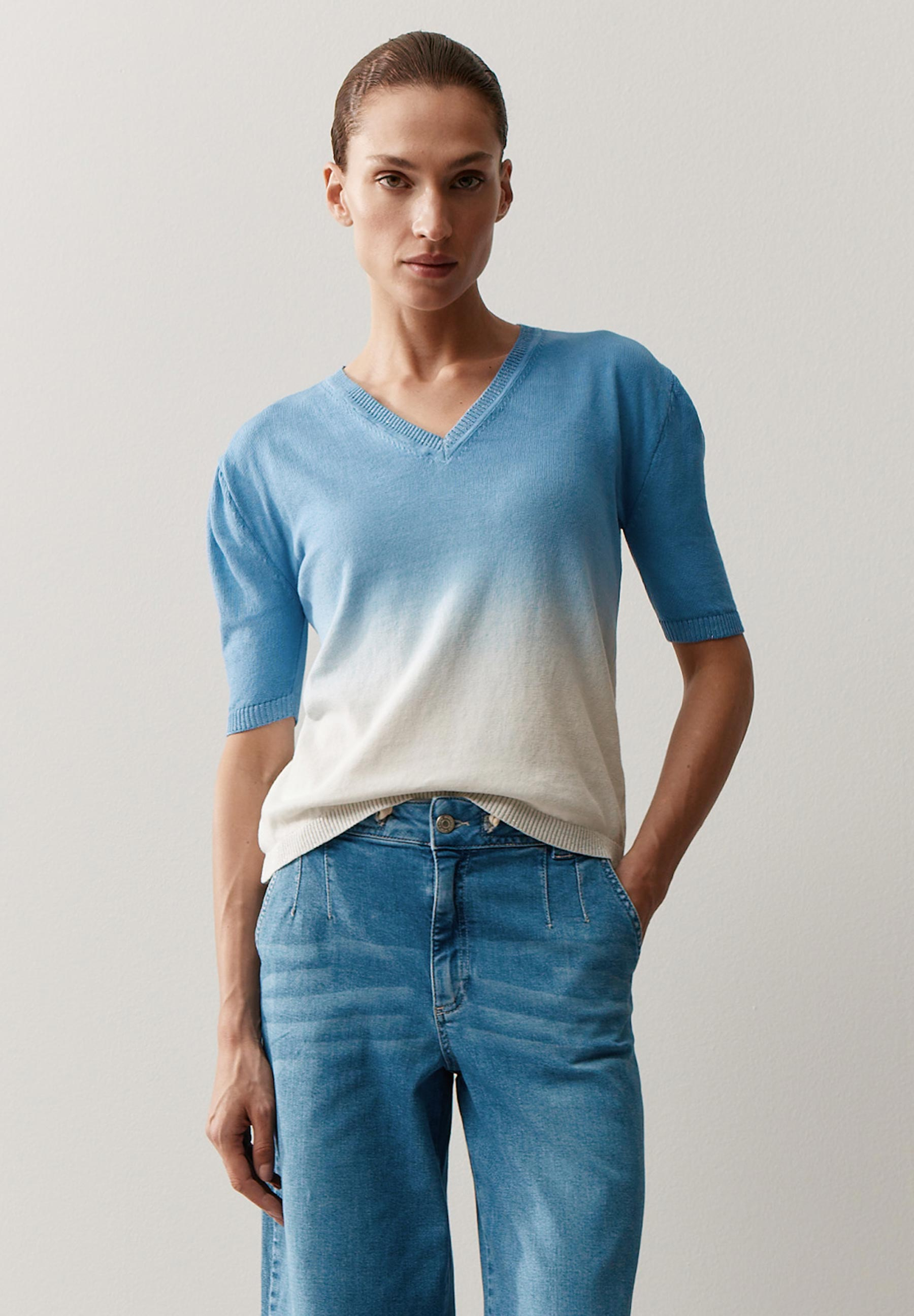 Eine Frau trägt ein blau-weißes MORE & MORE Oberteil mit V-Ausschnitt und Jeans vor einem hellen Hintergrund.