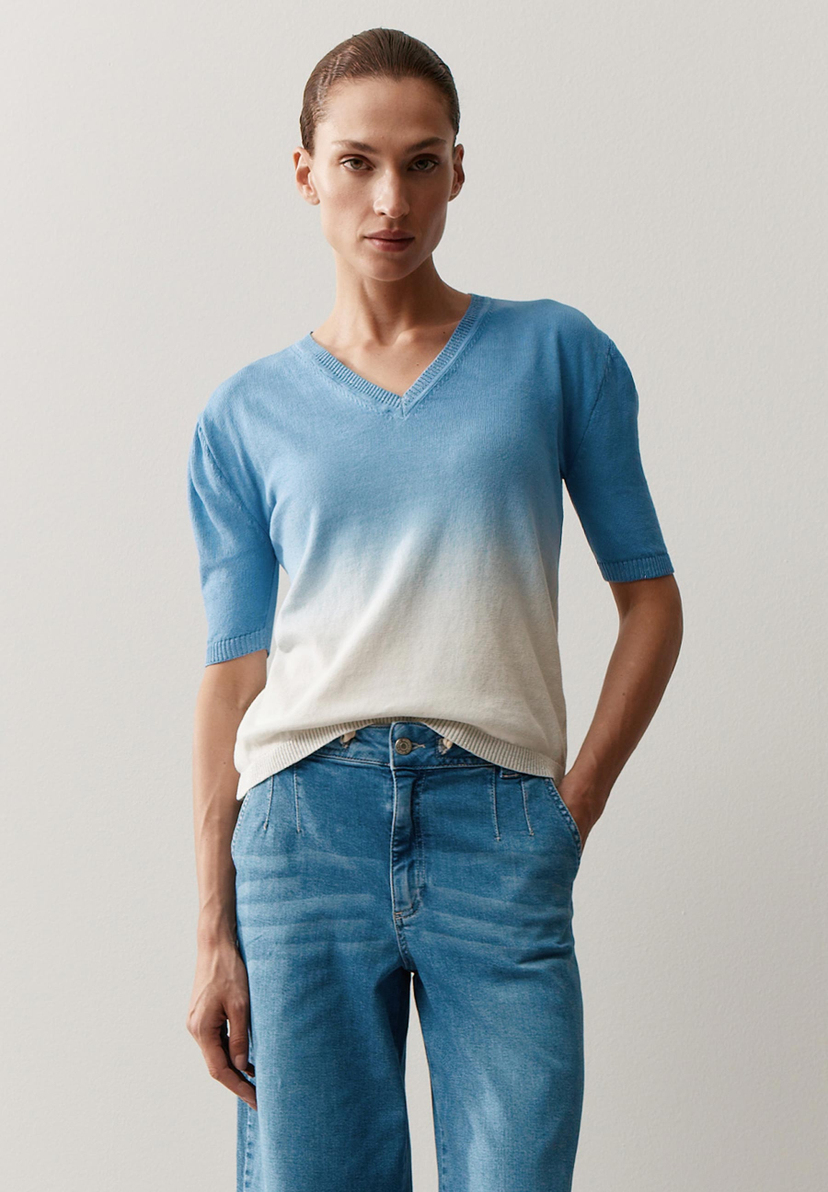Eine Frau trägt ein blau-weißes MORE & MORE Oberteil mit V-Ausschnitt und Jeans vor einem hellen Hintergrund. Eine Frau trägt ein blau-weißes MORE & MORE Oberteil mit V-Ausschnitt und Jeans vor einem hellen Hintergrund.