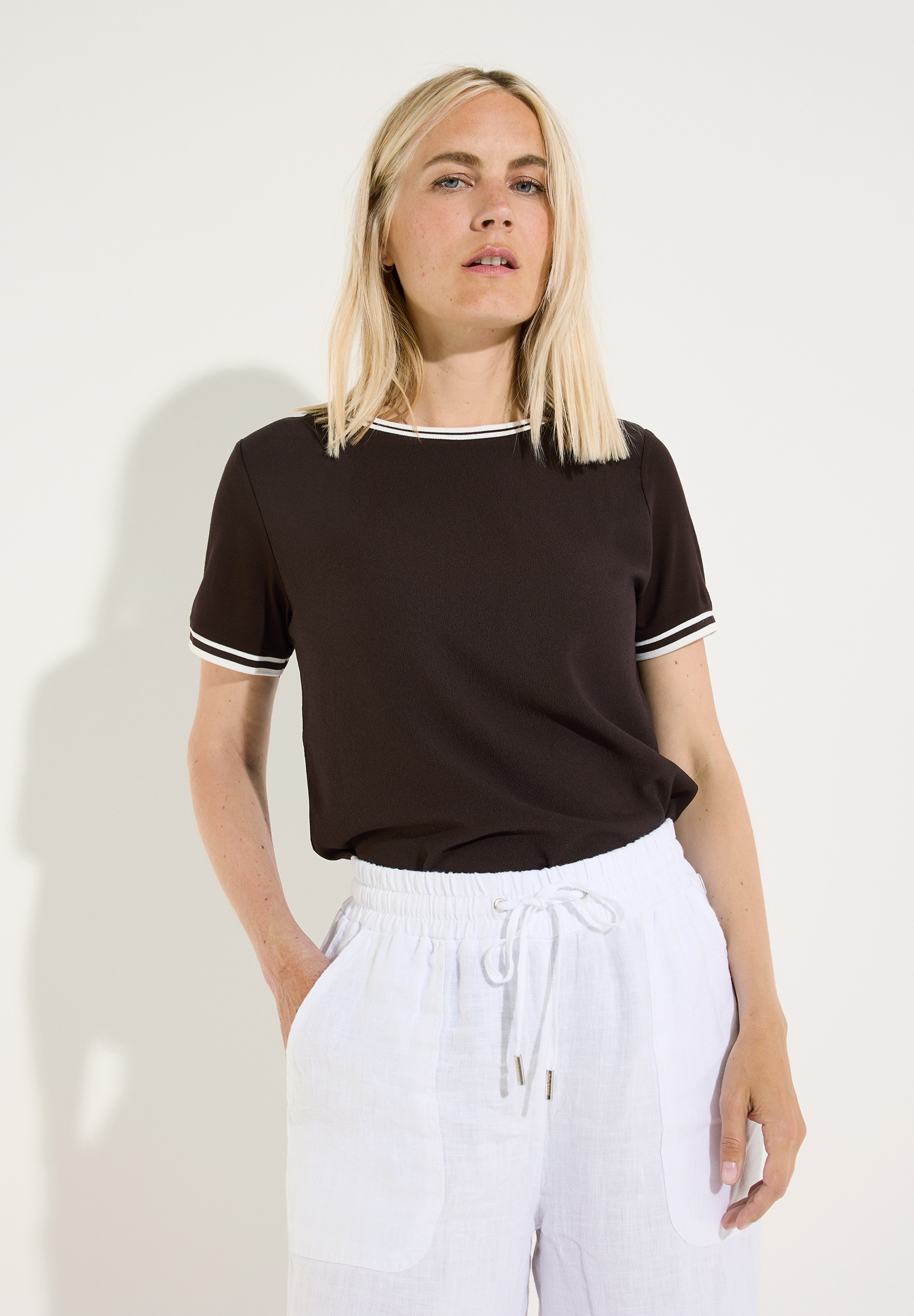 MORE & MORE Damenshirt, dunkelbraun, kurzärmlig mit hellen Streifen an Ärmeln und Hals, kombiniert mit weißer Leinenhose. MORE & MORE Damenshirt, dunkelbraun, kurzärmlig mit hellen Streifen an Ärmeln und Hals, kombiniert mit weißer Leinenhose.
