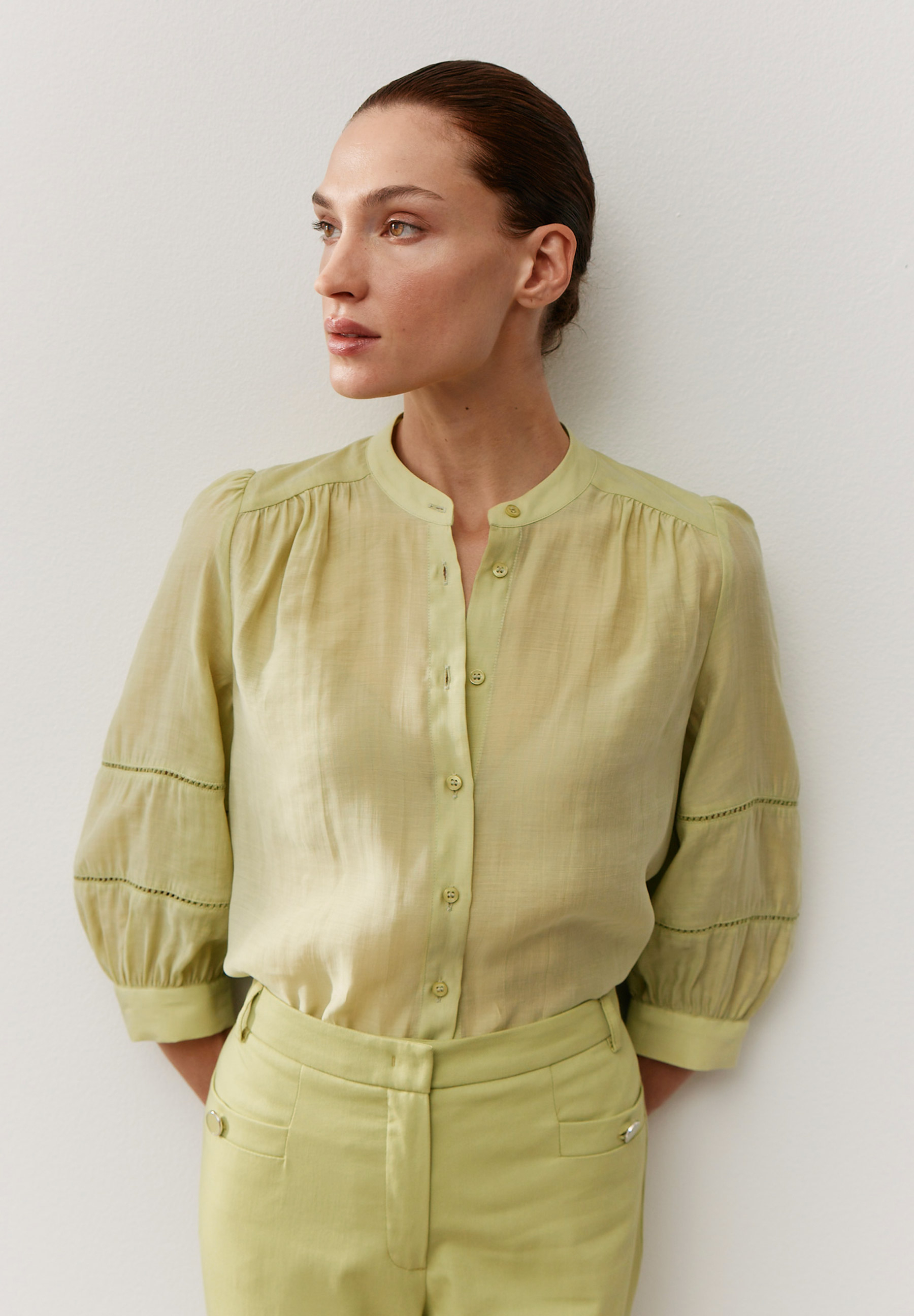 Eine Frau in einer hellgrünen MORE & MORE Bluse mit Webware und passender Hose vor einer weißen Wand.