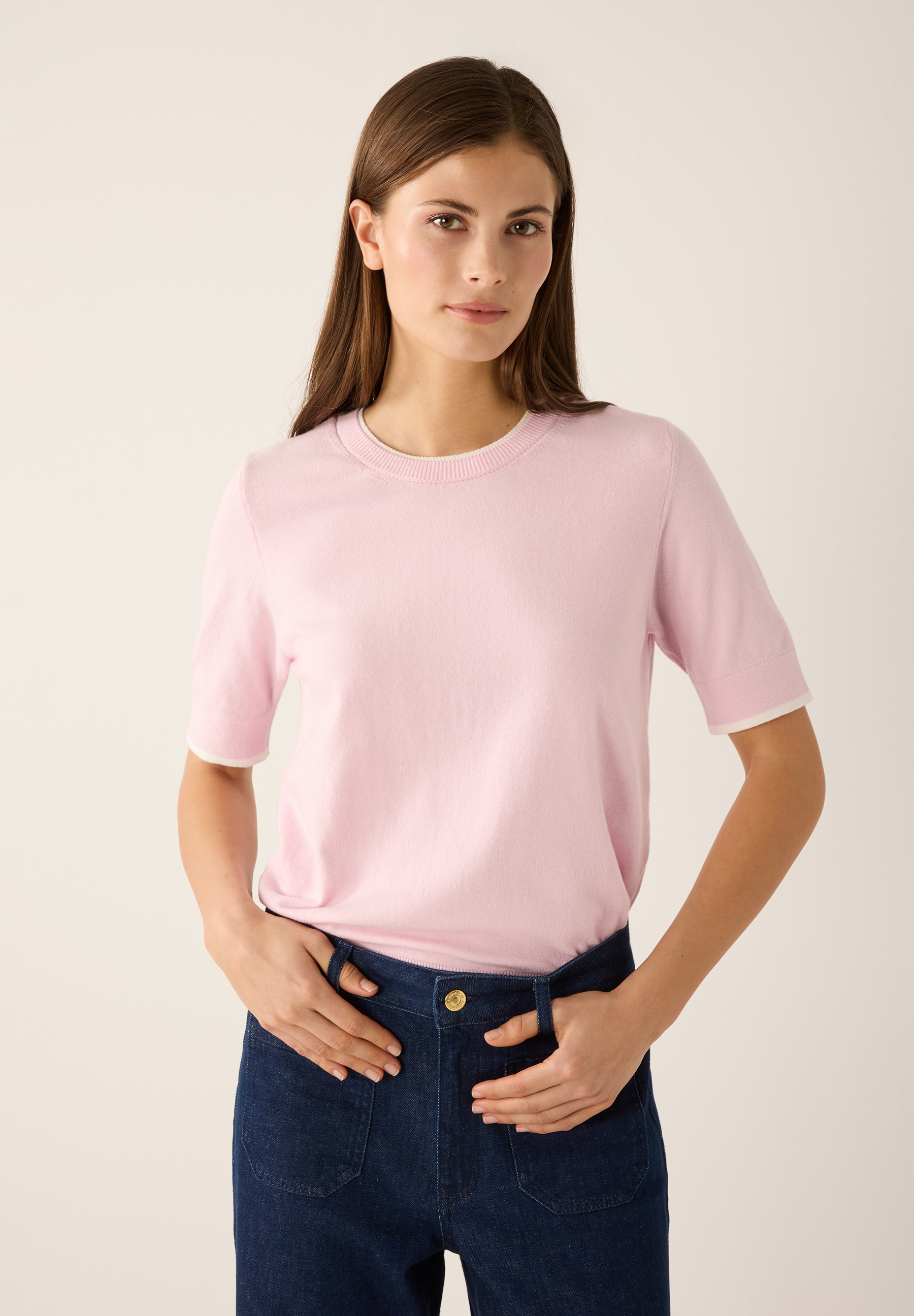 Eine Frau mit MORE & MORE Feinstrick-Oberteil. Der Pullover in Rosa hat kurze Ärmel mit weißen Kanten. Eine Frau mit MORE & MORE Feinstrick-Oberteil. Der Pullover in Rosa hat kurze Ärmel mit weißen Kanten.