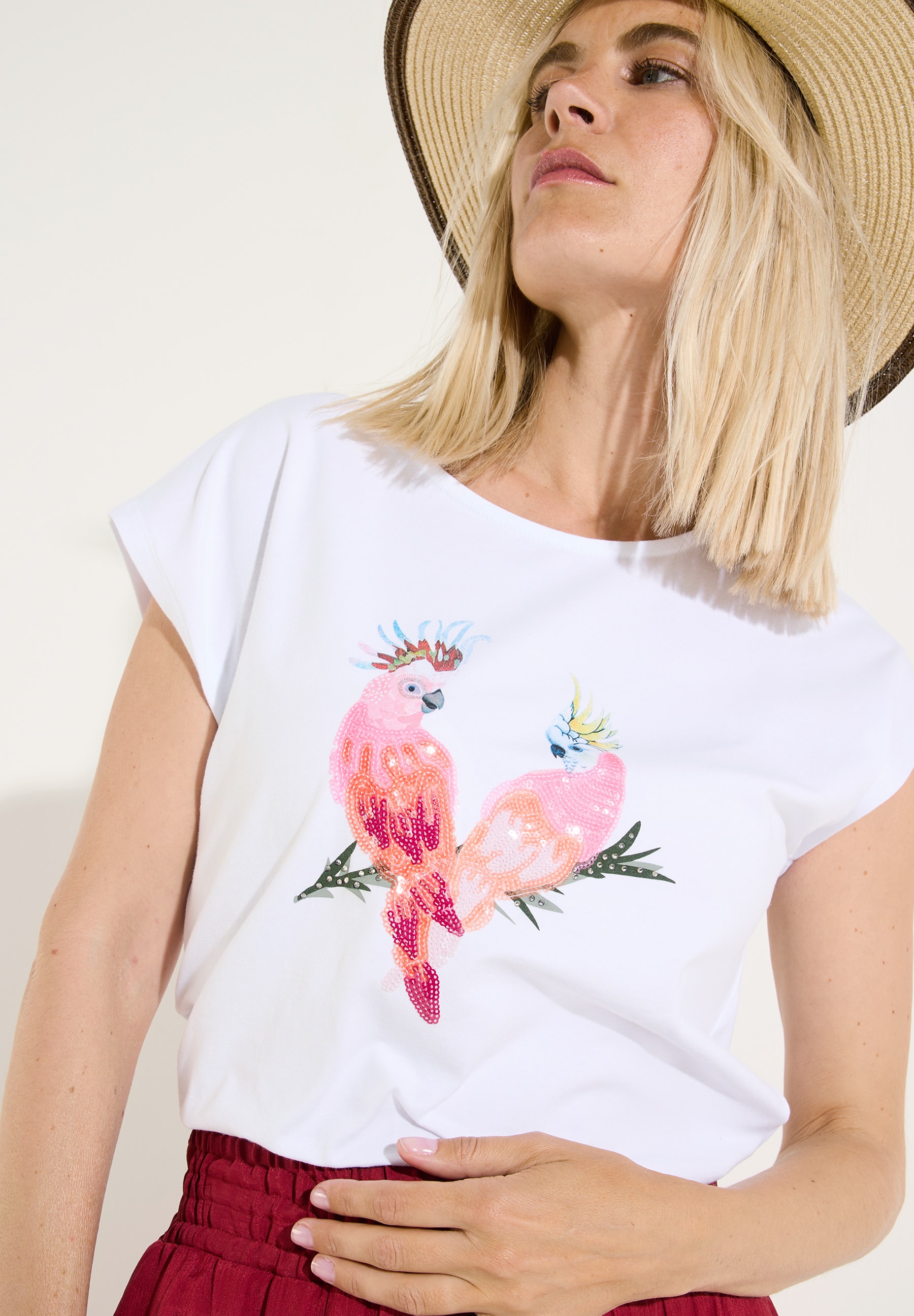 MORE & MORE T-Shirt/Top. Frau mit Hut trägt weißes Shirt mit zwei glitzernden Kakadus, die auf einem Zweig sitzen.