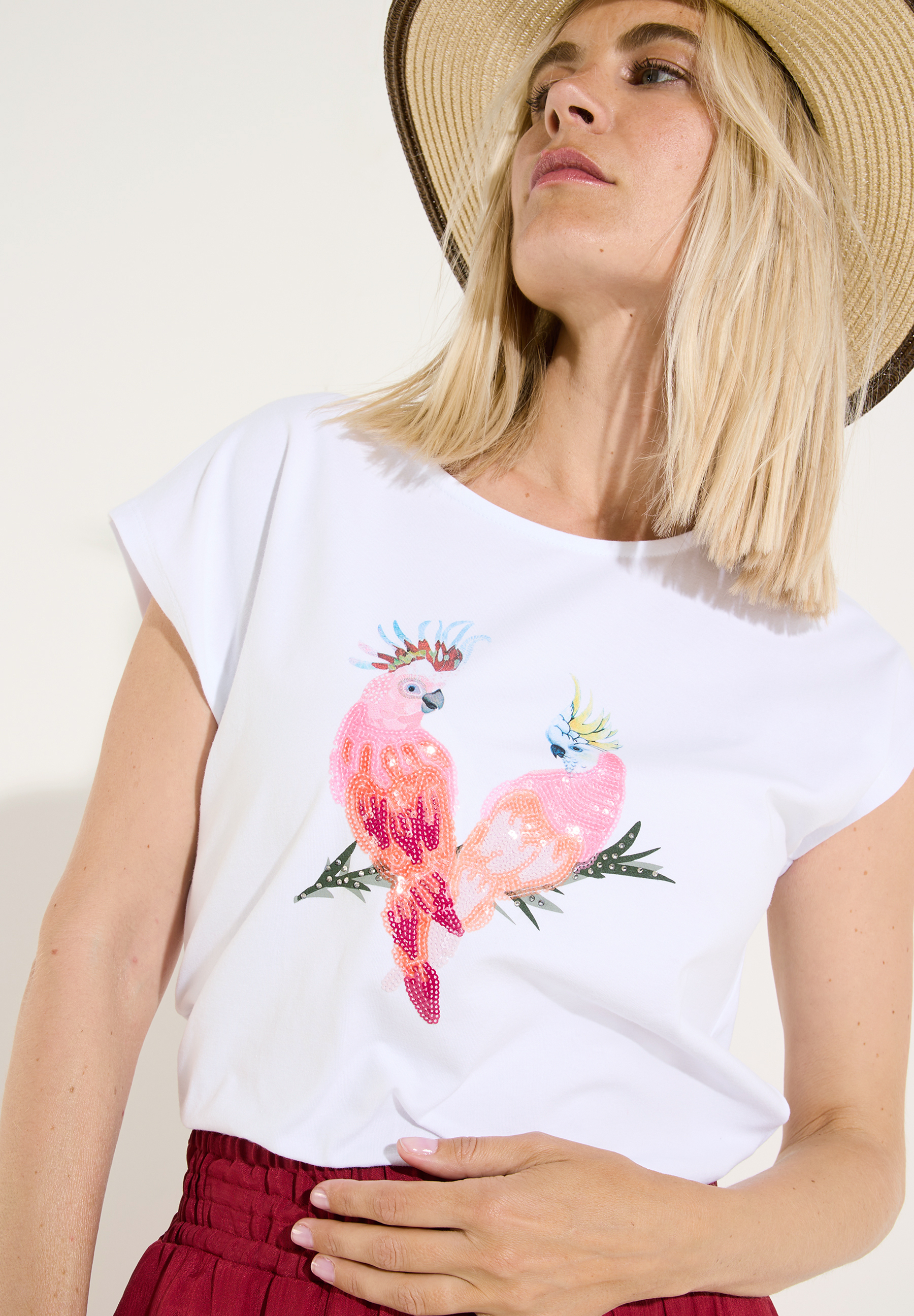 MORE & MORE T-Shirt/Top. Frau mit Hut trägt weißes Shirt mit zwei glitzernden Kakadus, die auf einem Zweig sitzen.