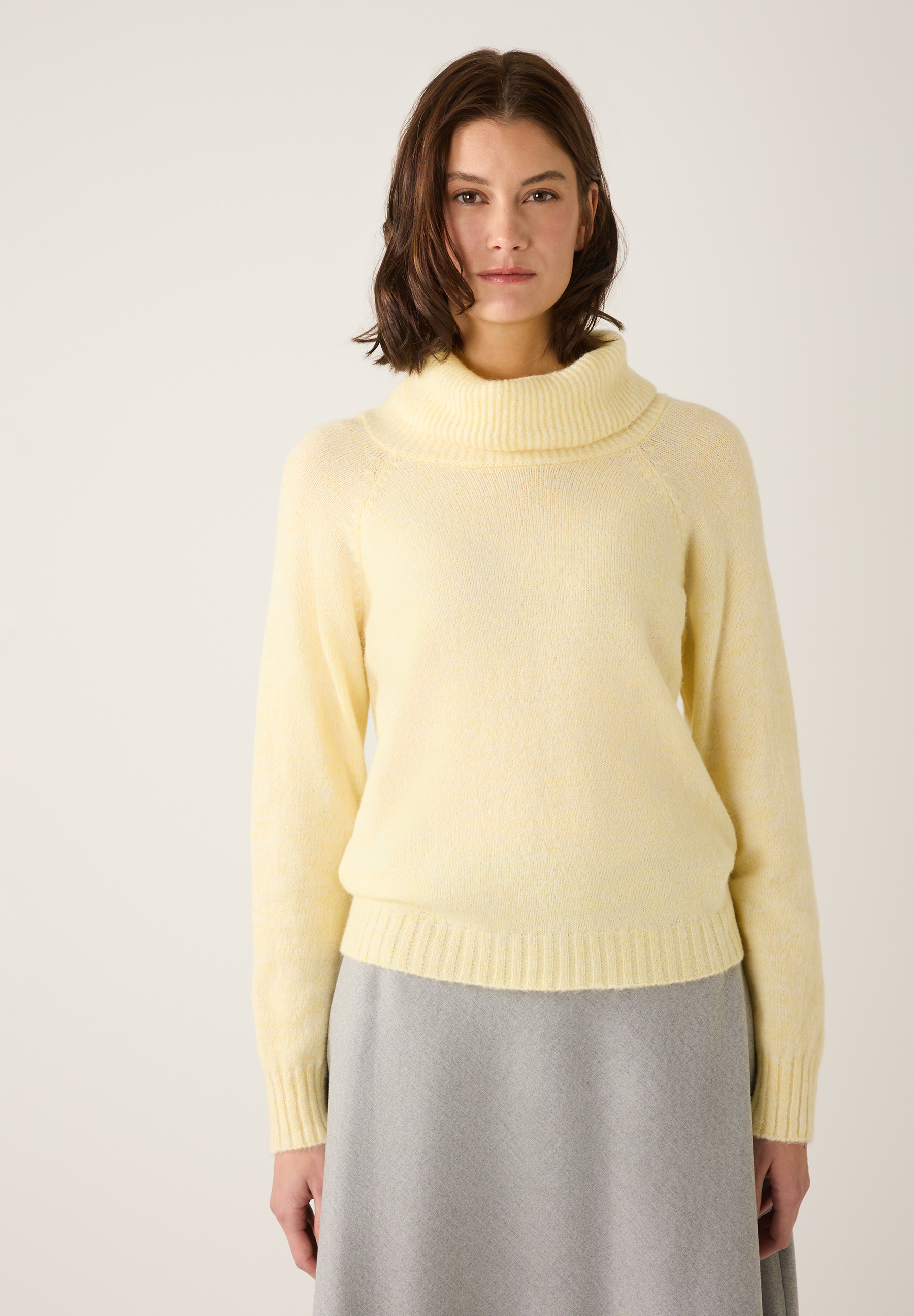 Eine Frau trägt einen hellgelben Strickpullover von MORE & MORE mit Rollkragen und einen grauen Rock.