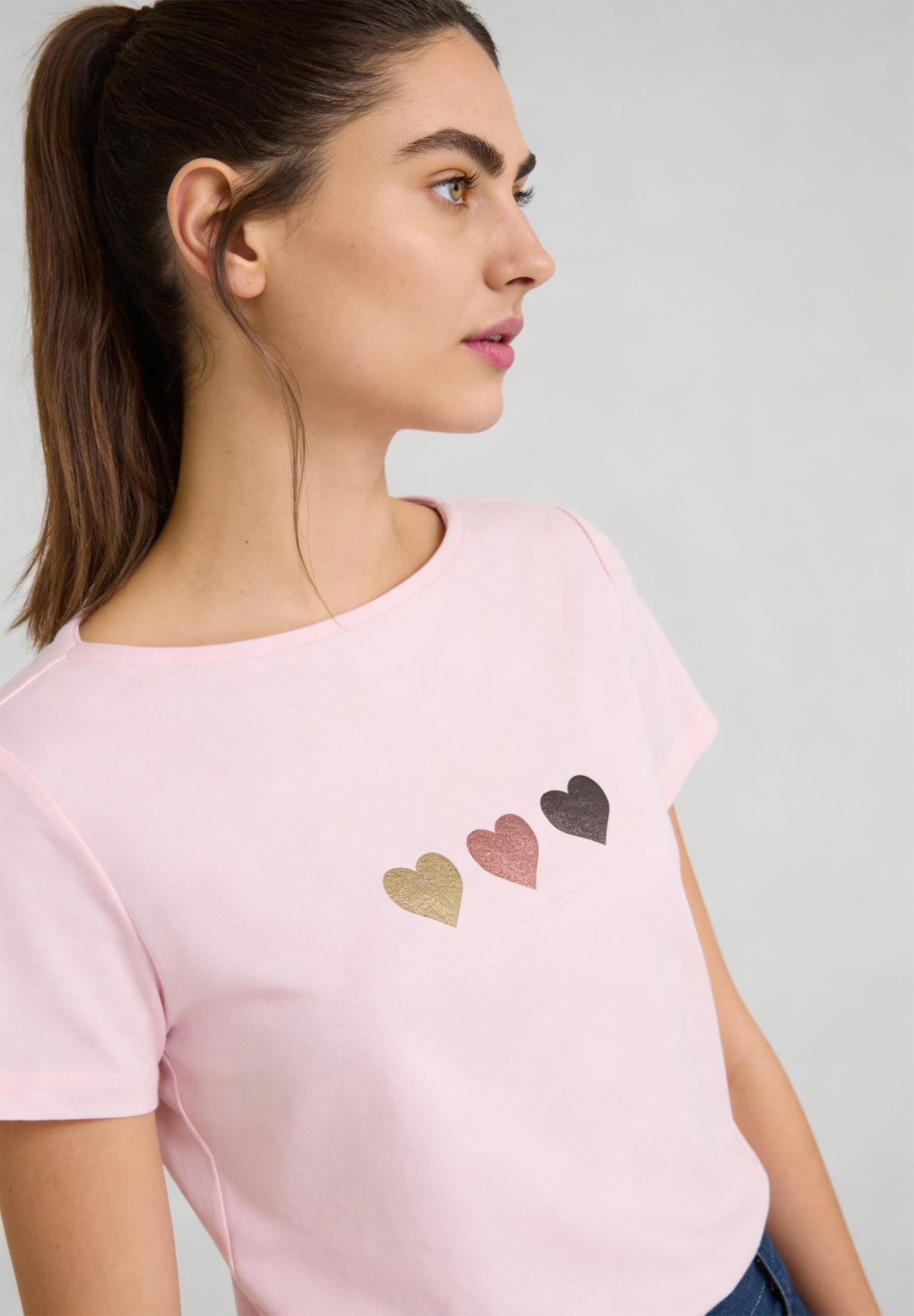 Profilansicht einer Frau mit MORE & MORE Oberteil. Das T-Shirt ist rosa und mit drei Herzen verziert.