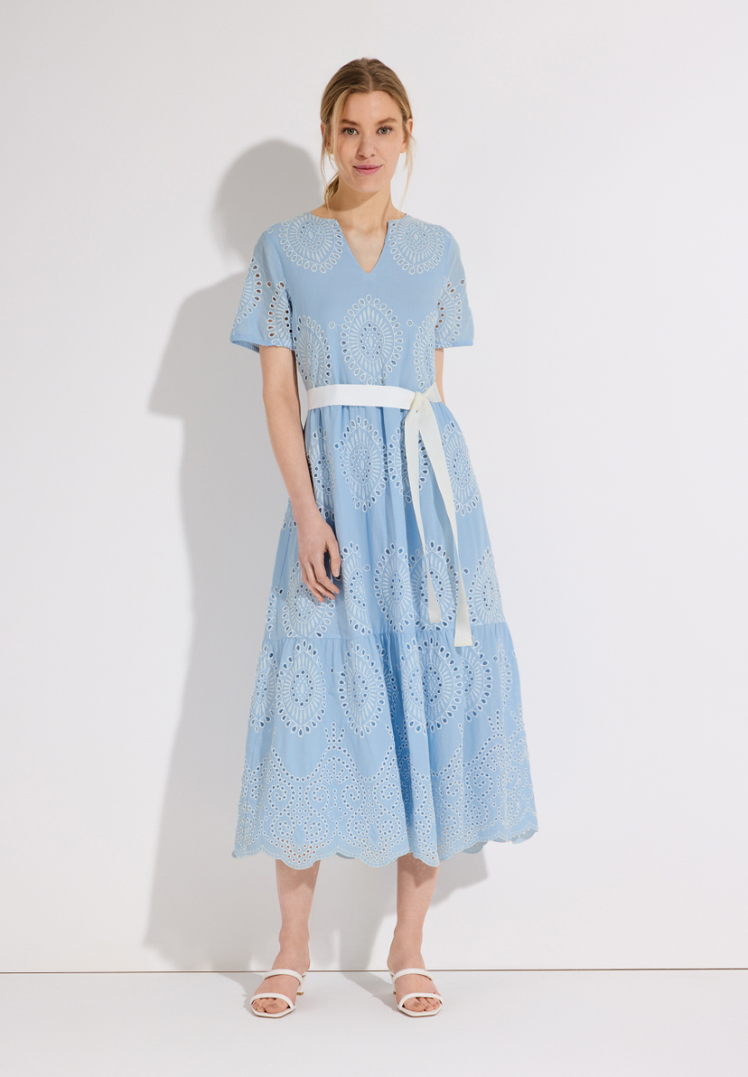 Eine Frau in einem hellblauen MORE & MORE Oberteil mit Lochstickerei und weißem Gürtel. Eine Frau in einem hellblauen MORE & MORE Oberteil mit Lochstickerei und weißem Gürtel.