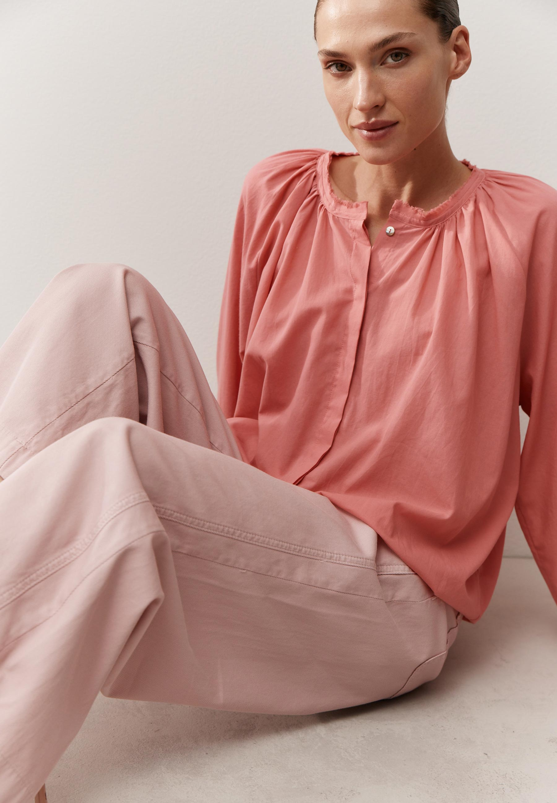 Eine Frau mit einer rosafarbenen MORE & MORE Bluse und passender Hose sitzt vor einer hellen Wand.