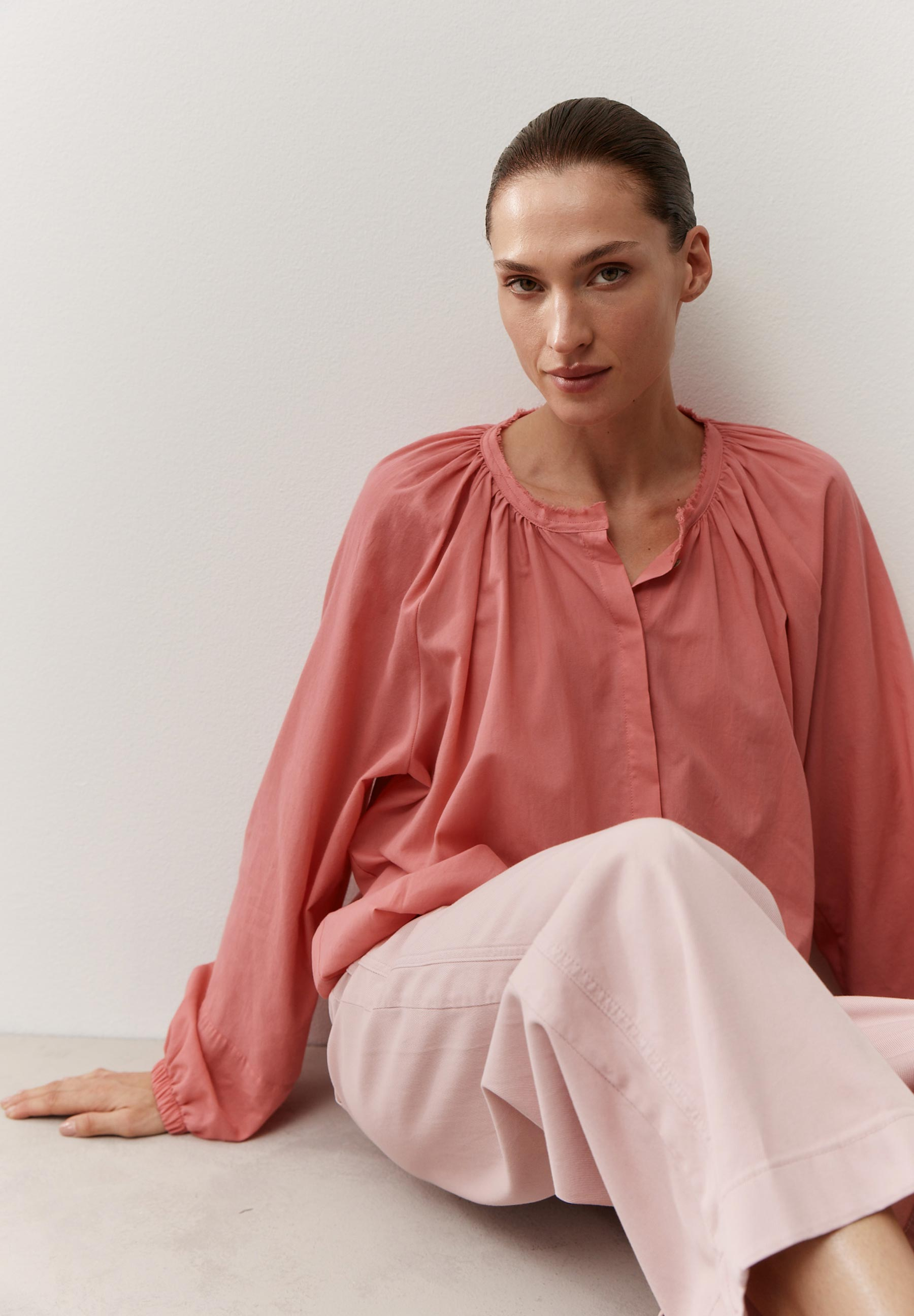 Eine Frau in MORE & MORE Bluse und Hose, lehnt an einer weißen Wand.