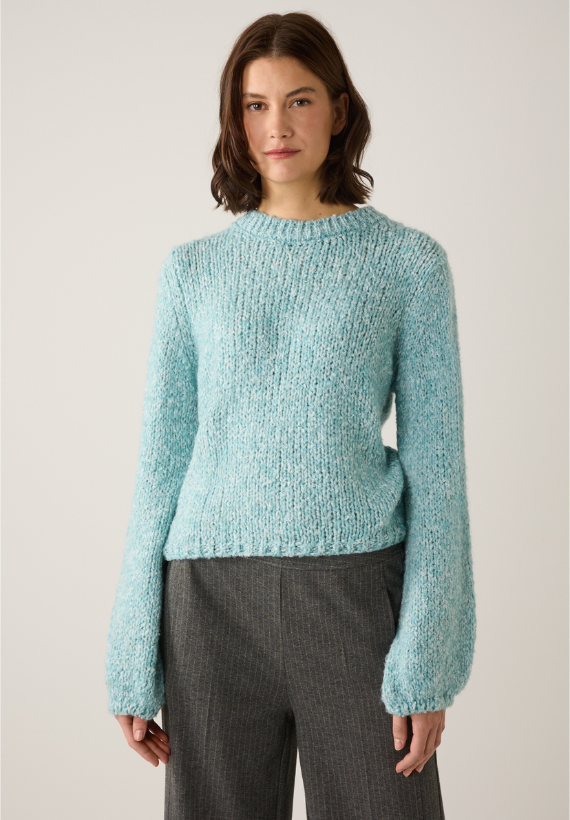 Eine Frau trägt einen hellblauen Grobstrickpullover der Marke MORE & MORE mit grau gestreiften Hosen. Eine Frau trägt einen hellblauen Grobstrickpullover der Marke MORE & MORE mit grau gestreiften Hosen.