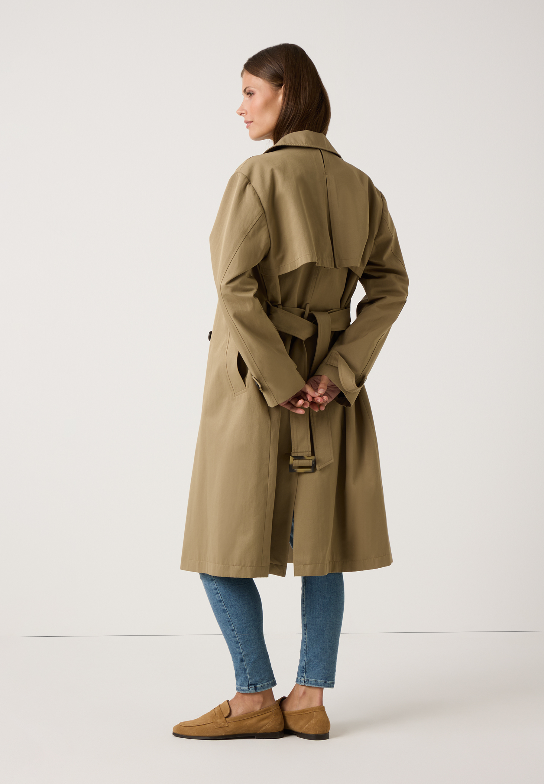 Eine Frau von hinten in einem MORE & MORE Trenchcoat aus Gabardine, kombiniert mit Jeans und Loafern. Eine Frau von hinten in einem MORE & MORE Trenchcoat aus Gabardine, kombiniert mit Jeans und Loafern.