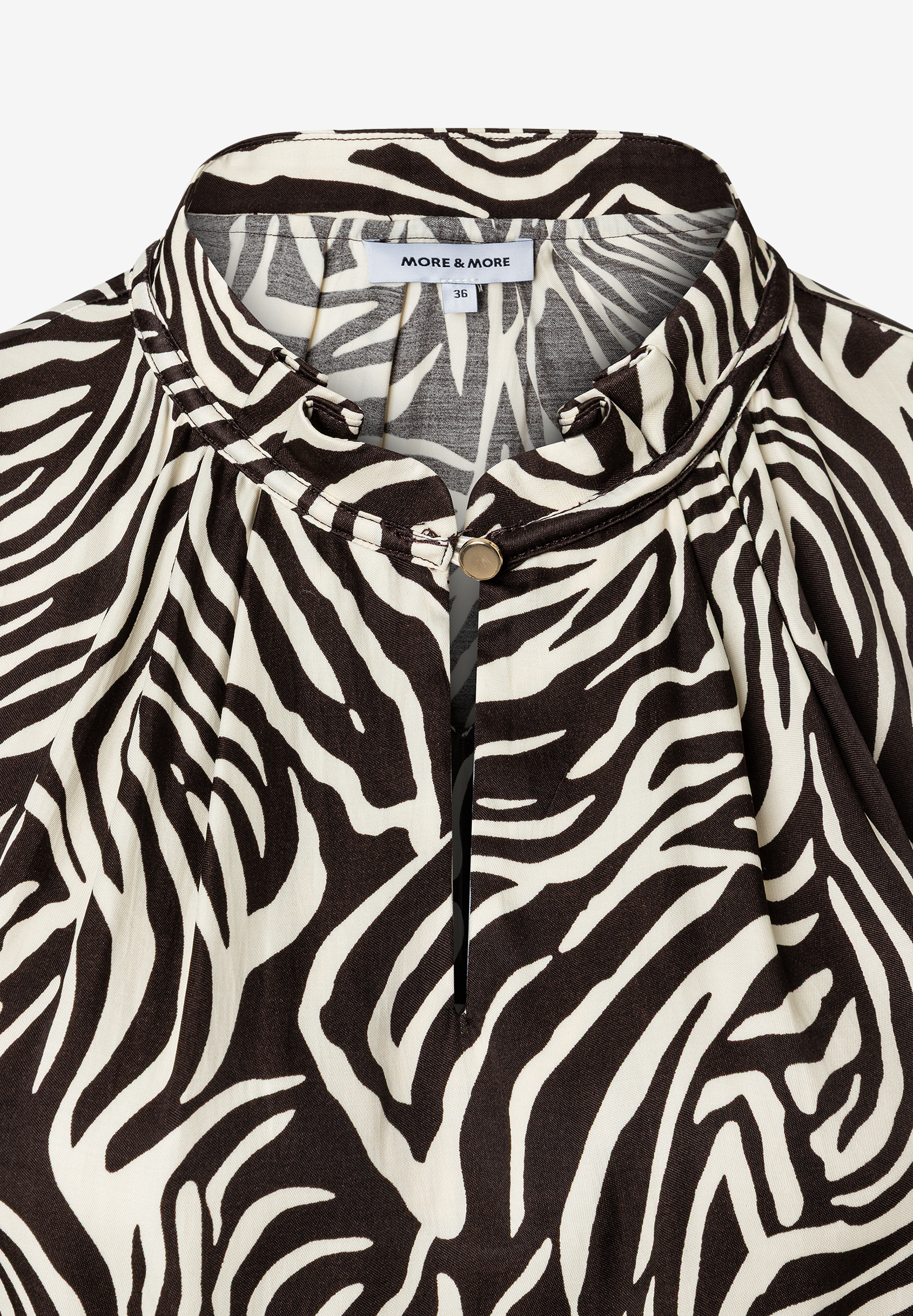 Nahaufnahme einer MORE & MORE Bluse mit Zebramuster aus fließender Viskose, Knopf am Kragen und Etikett. Nahaufnahme einer MORE & MORE Bluse mit Zebramuster aus fließender Viskose, Knopf am Kragen und Etikett.