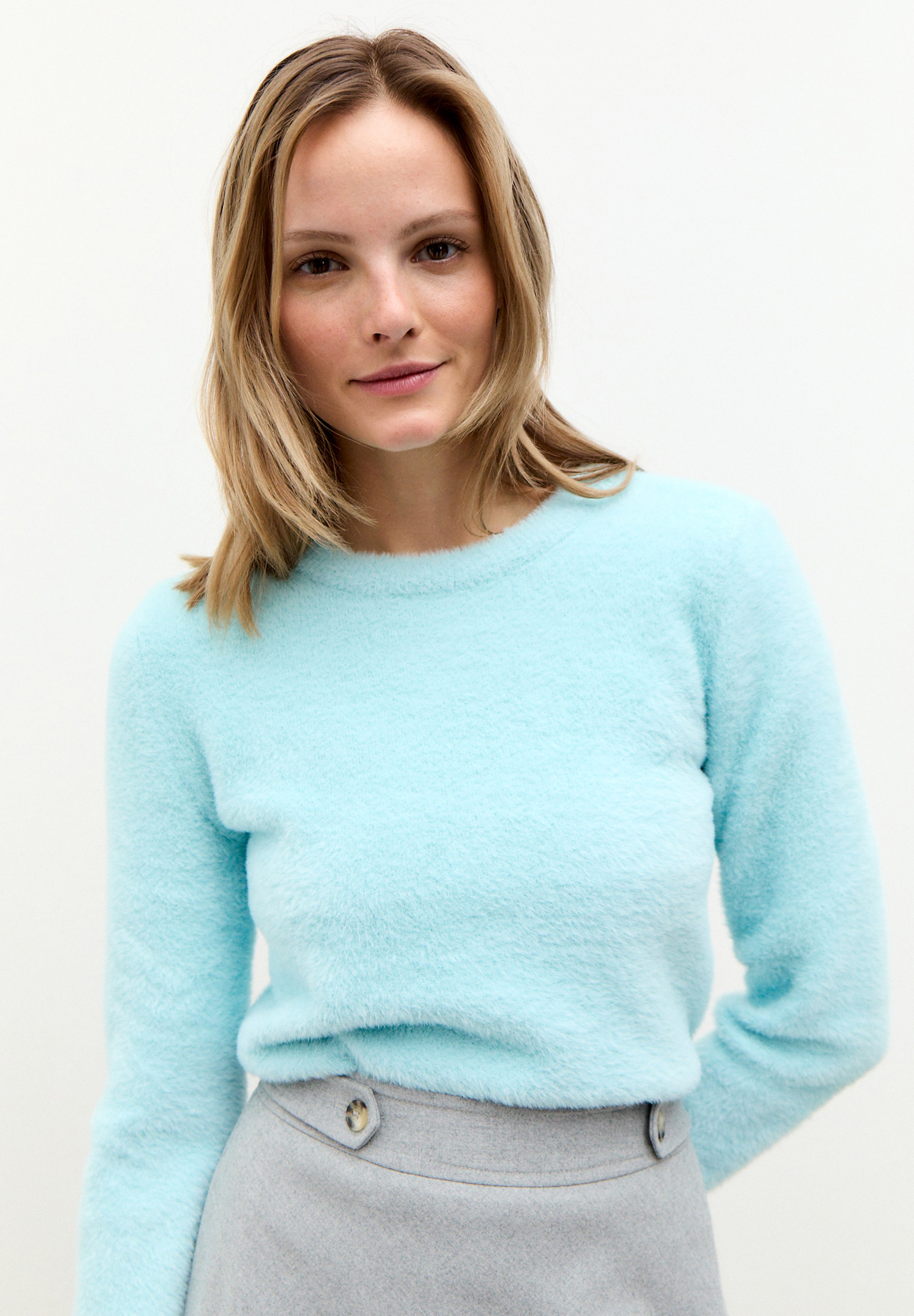 Eine Frau trägt einen hellblauen MORE & MORE Strickpullover mit weichem Griff und einen grauen Rock.