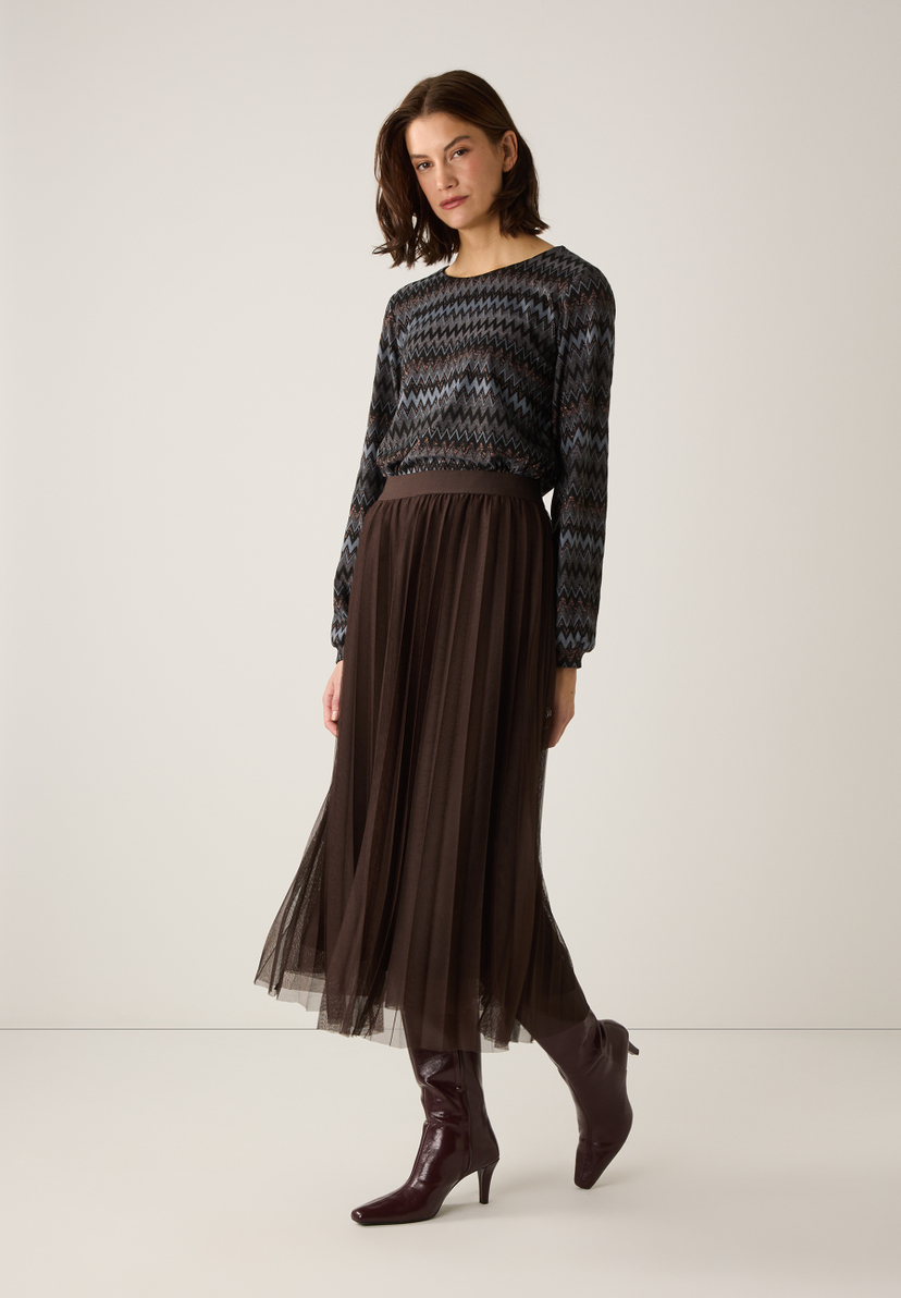 Eine Frau trägt ein MORE & MORE Oberteil mit Zickzackmuster, einen Faltenrock und Stiefel vor einem hellen Hintergrund. Eine Frau trägt ein MORE & MORE Oberteil mit Zickzackmuster, einen Faltenrock und Stiefel vor einem hellen Hintergrund.
