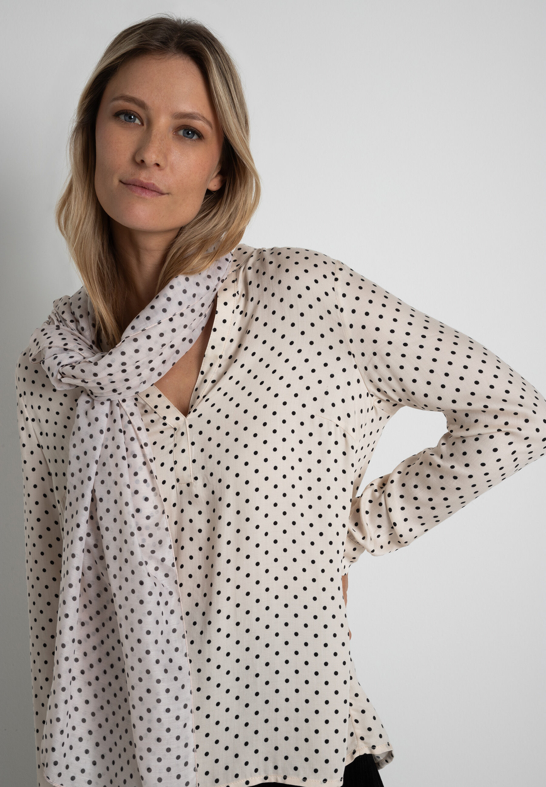 Eine Frau mit blonden, gewellten Haaren trägt ein gepunktetes Hemd und einen passenden Schal von MORE & MORE Accessoires.