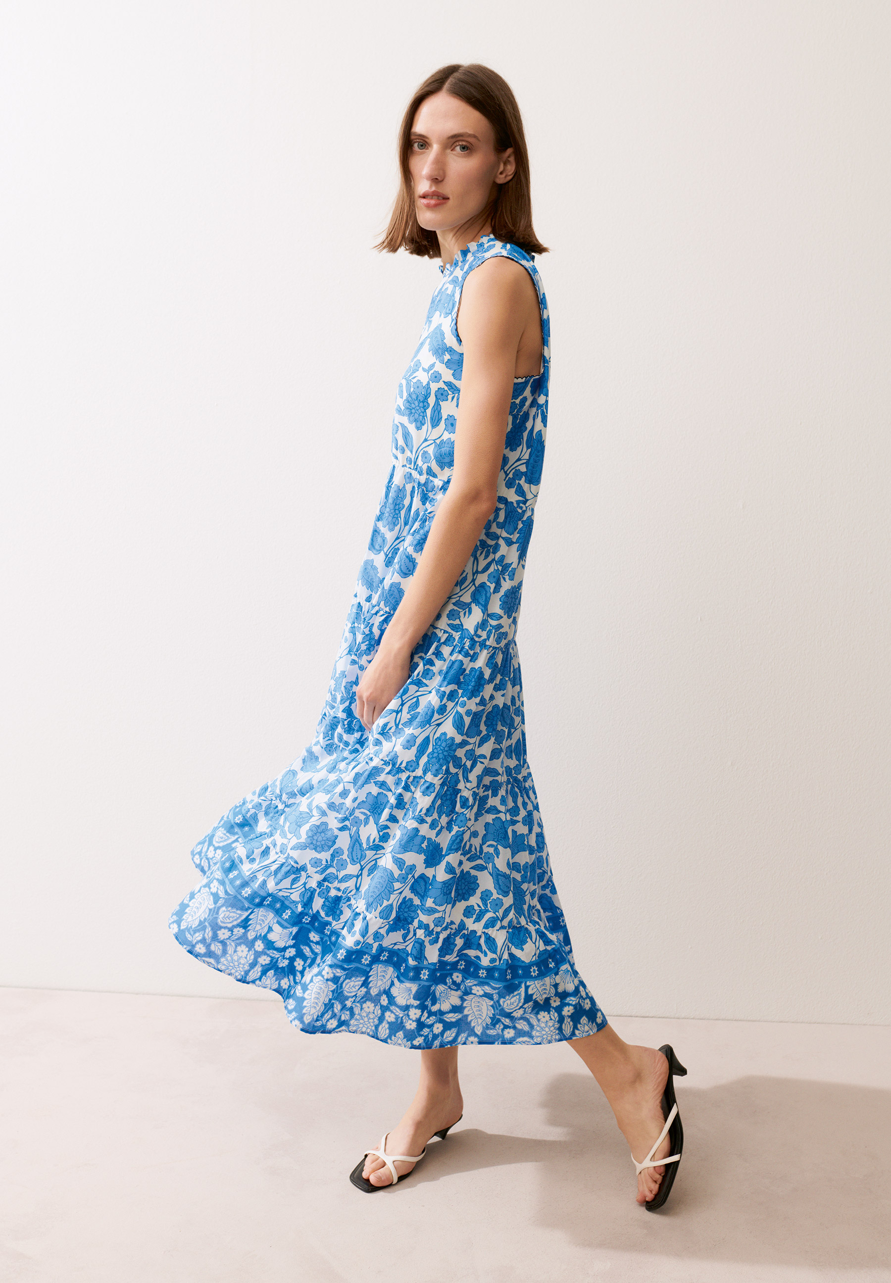 Eine Frau, die ein blau-weißes MORE & MORE Kleid mit Blumenmuster und Riemchensandalen trägt.