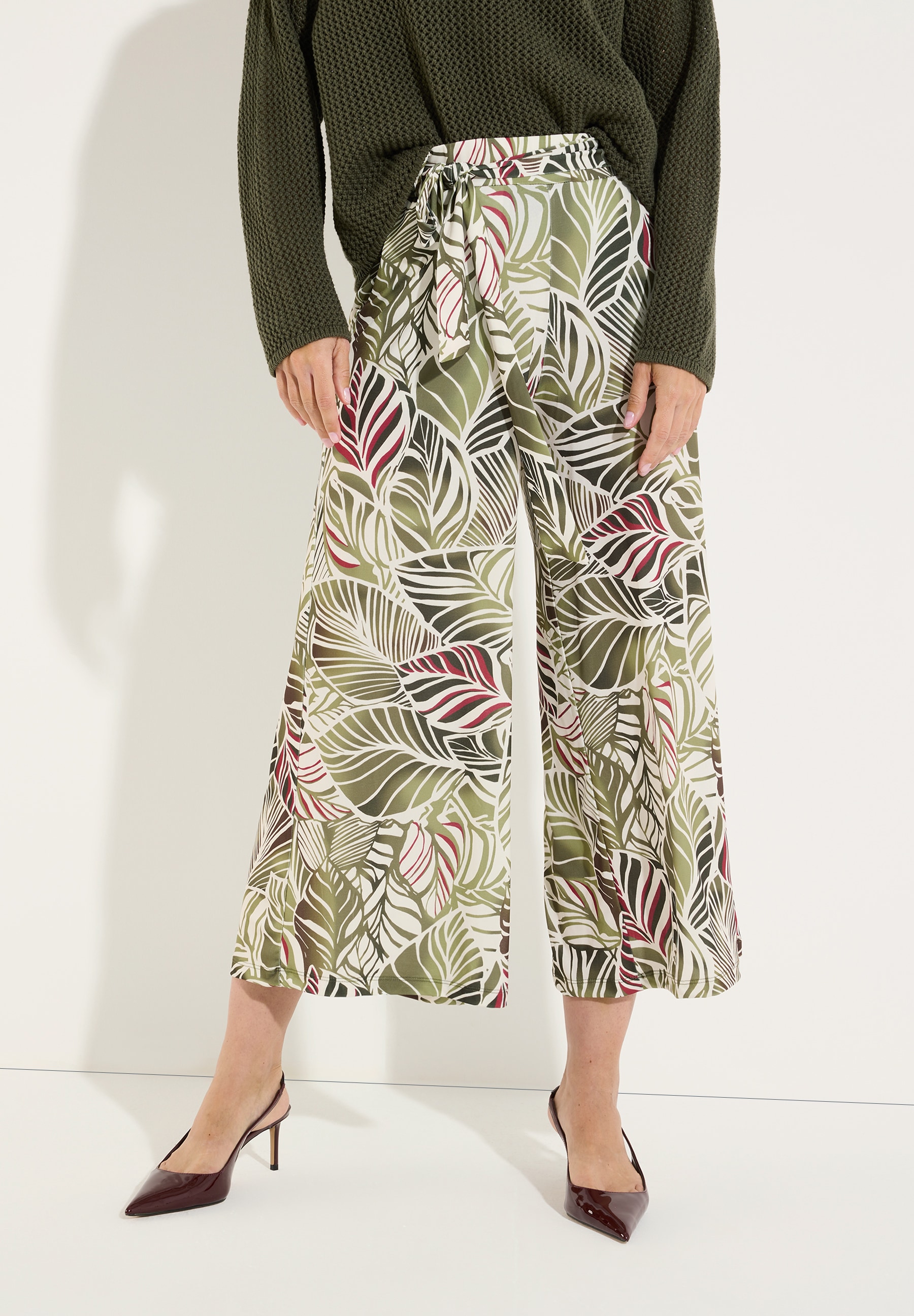 MORE & MORE Jersey-Hose mit Palmenmuster in Grün, Weiß & Rot, kombiniert mit weißen Sandalen.
