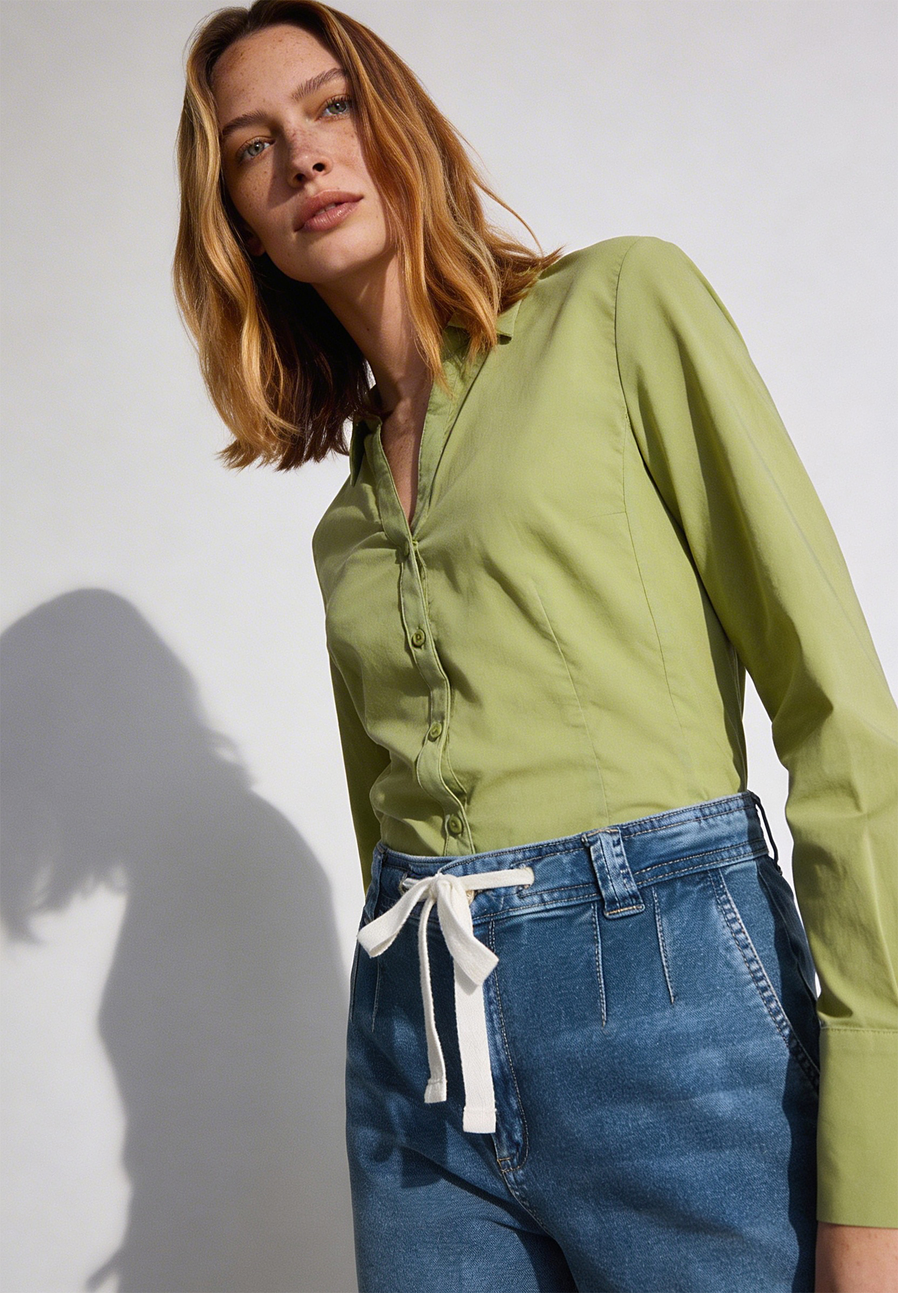 Eine Frau mit Sommersprossen trägt eine grüne MORE & MORE Popeline-Bluse und Jeans. Eine Frau mit Sommersprossen trägt eine grüne MORE & MORE Popeline-Bluse und Jeans.