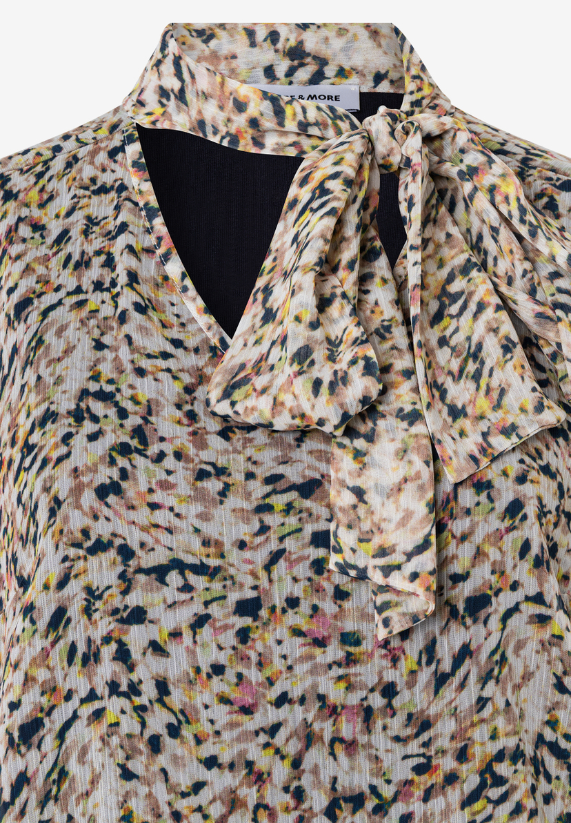 Nahaufnahme einer bedruckten MORE & MORE Bluse mit Schleifendetail am Ausschnitt. Nahaufnahme einer bedruckten MORE & MORE Bluse mit Schleifendetail am Ausschnitt.