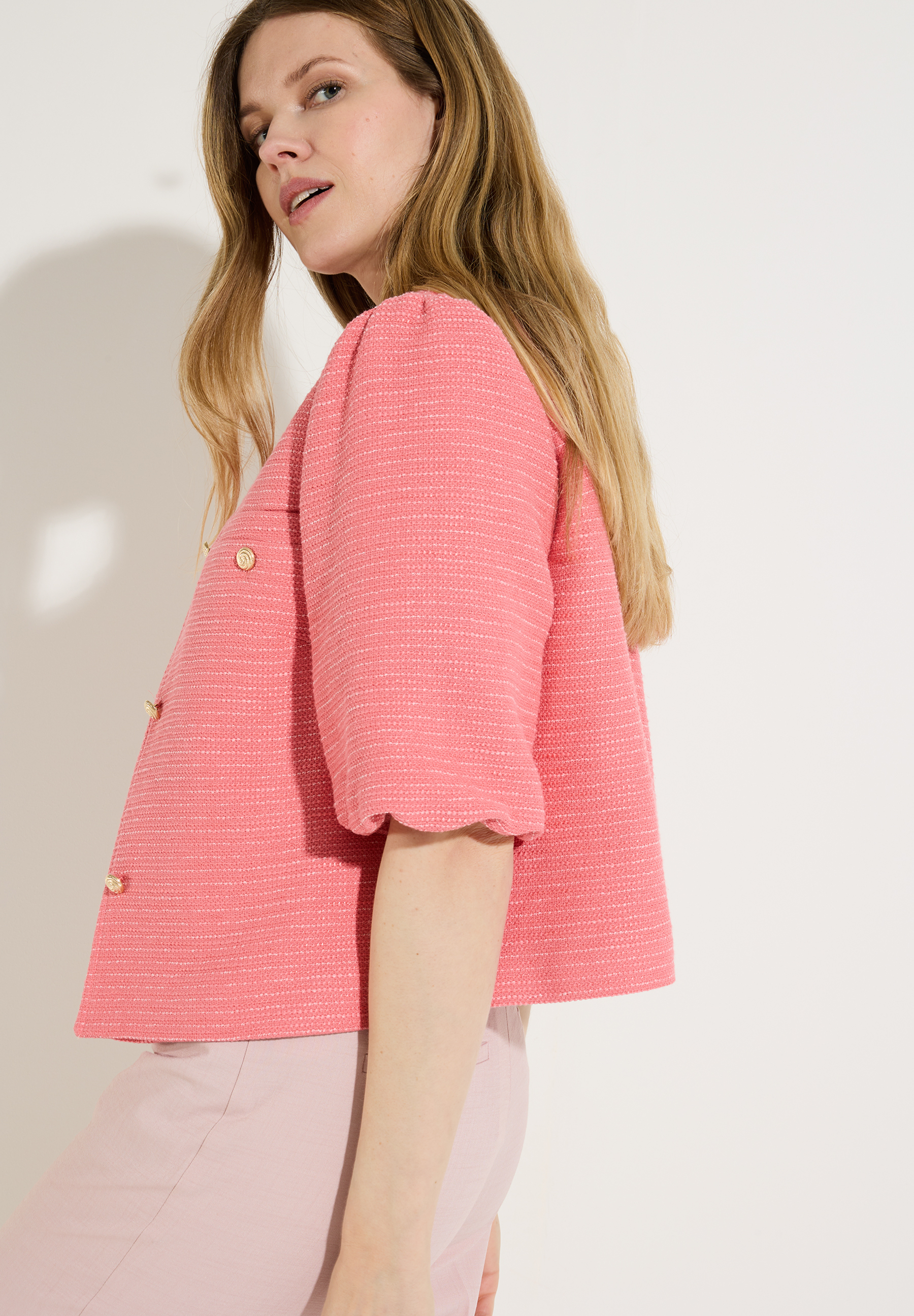 Eine Frau präsentiert einen lachsfarbenen Bouclé-Blazer von MORE & MORE mit goldenen Knöpfen.