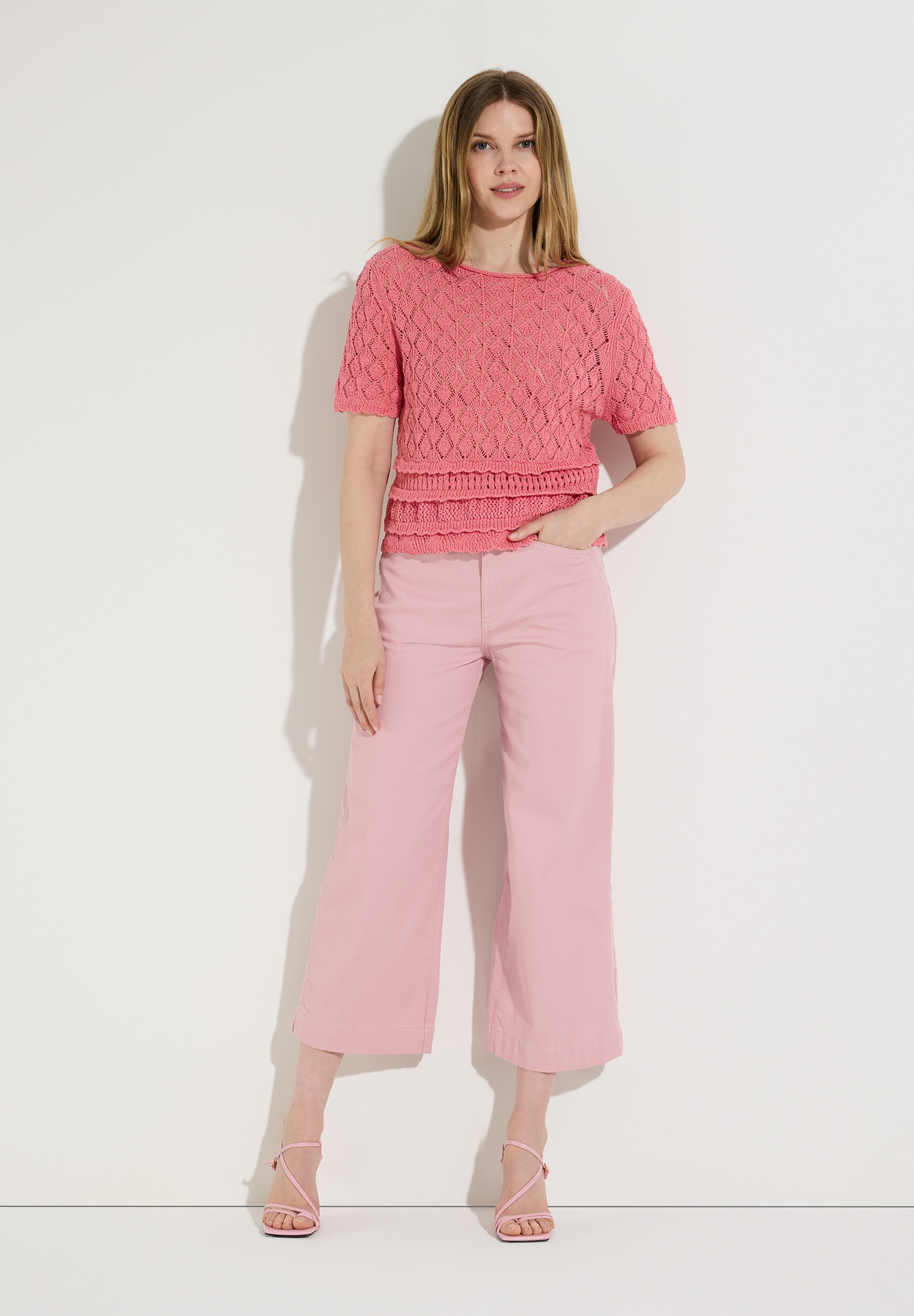Eine Frau in einem rosafarbenen MORE & MORE Strickoberteil mit Lochmuster und einer passenden Hose vor einer weißen Wand.