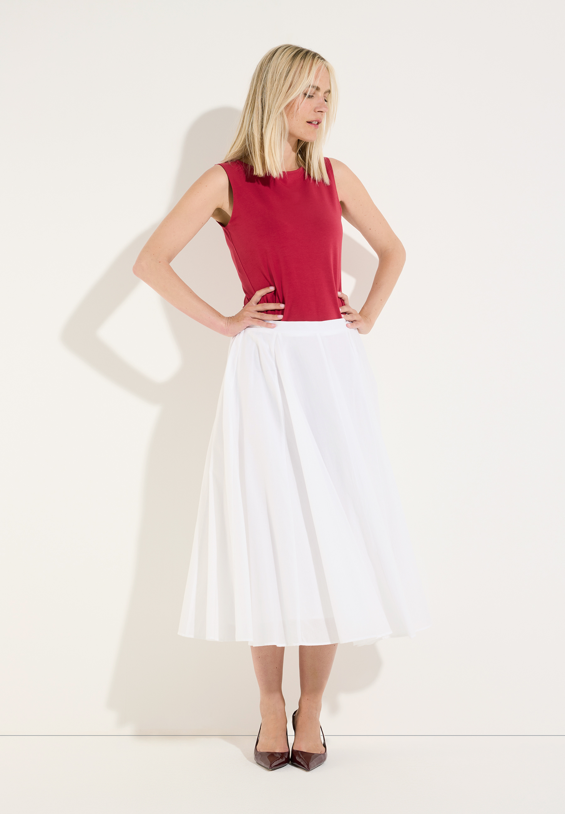 MORE & MORE Jersey-Top in Rot kombiniert mit weißem Midirock und braunen Pumps. Frau mit den Händen in den Hüften. MORE & MORE Jersey-Top in Rot kombiniert mit weißem Midirock und braunen Pumps. Frau mit den Händen in den Hüften.