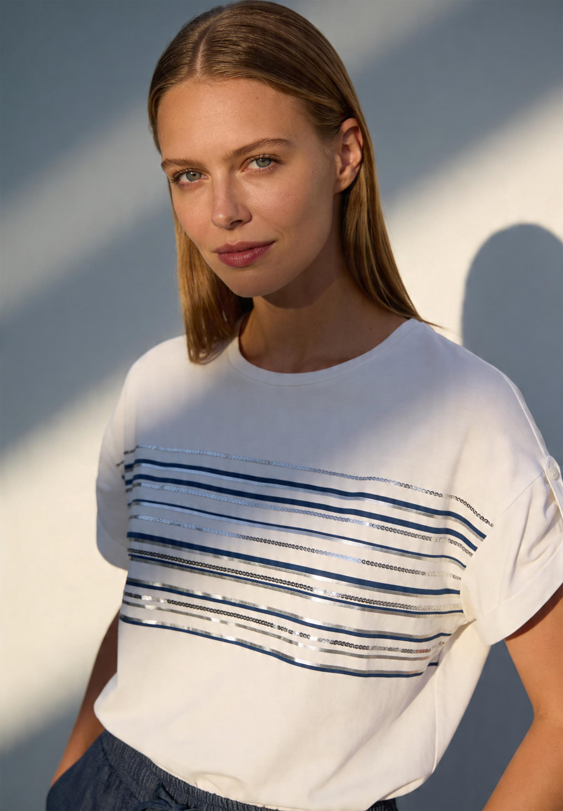 Porträt einer Frau mit MORE & MORE T-Shirt mit blau-silbernen Streifen vor hellem Hintergrund. Porträt einer Frau mit MORE & MORE T-Shirt mit blau-silbernen Streifen vor hellem Hintergrund.
