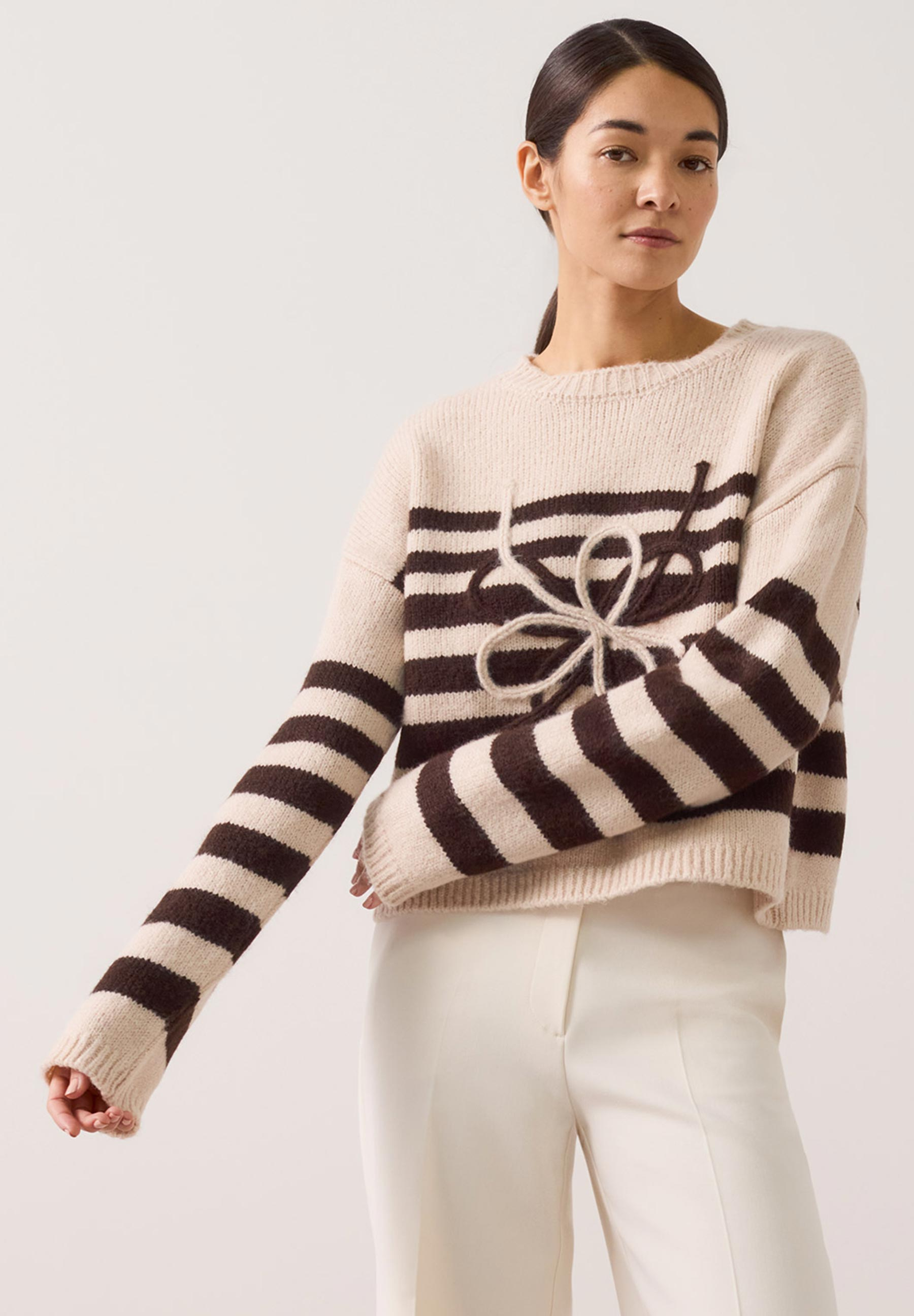 MORE & MORE Pullover: Gestreifter Strickpullover in Creme & Braun mit Schleifendetail. Eine Frau trägt ihn zu weißer Hose.