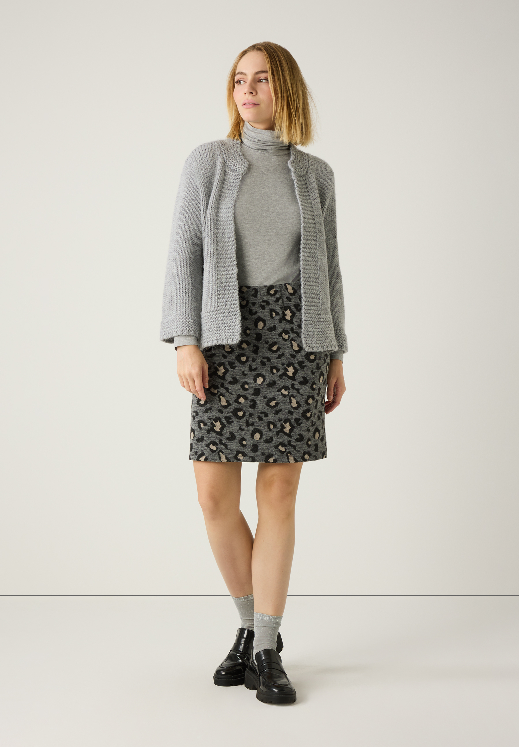 Eine Frau in einem MORE & MORE Oberteil mit Rollkragen, Strickjacke und Rock mit Tiermuster vor einem weißen Hintergrund. Eine Frau in einem MORE & MORE Oberteil mit Rollkragen, Strickjacke und Rock mit Tiermuster vor einem weißen Hintergrund.