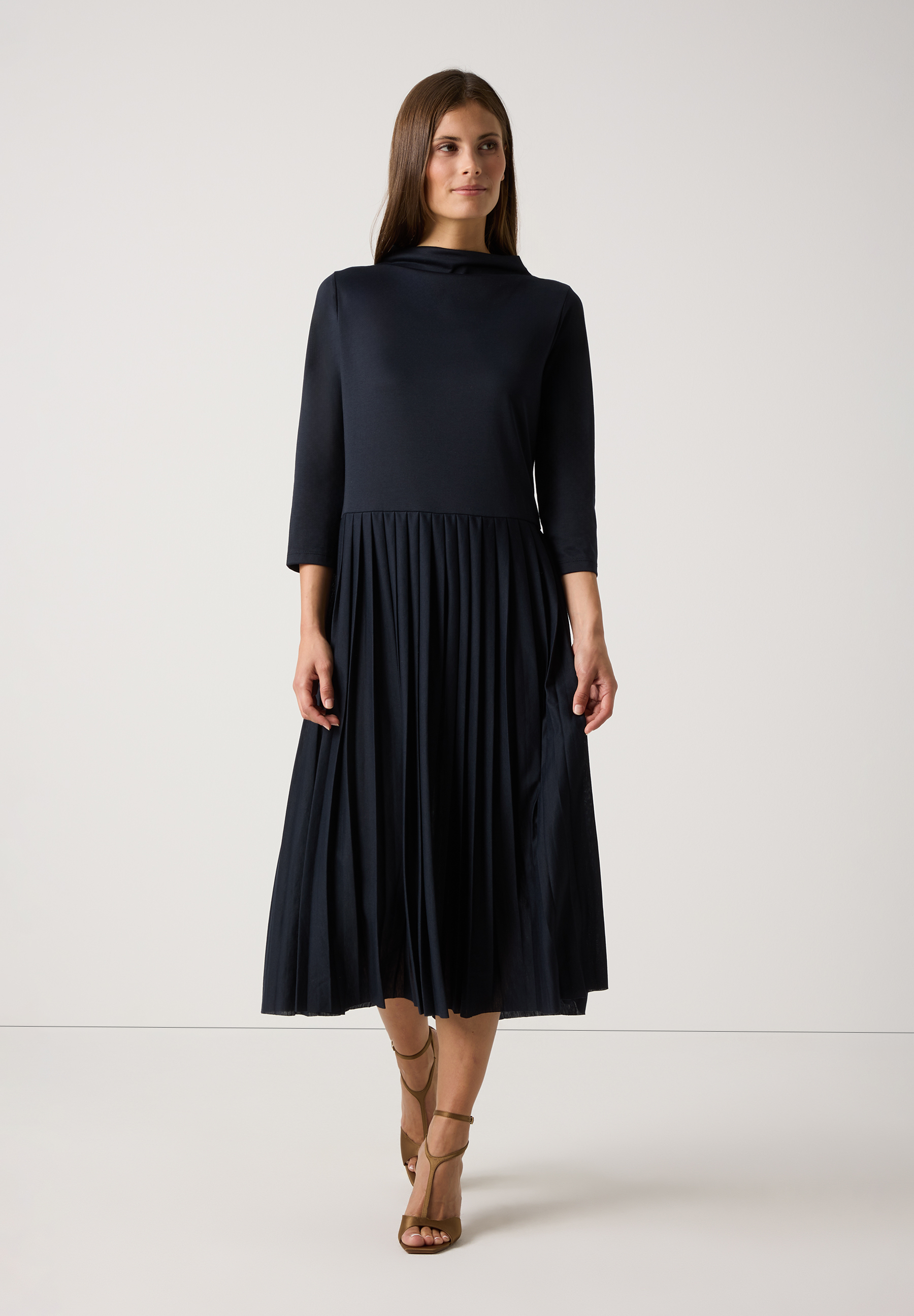 Eine Frau in einem dunkelblauen MORE & MORE Jersey-Kleid mit 3/4-Ärmeln und Faltenrock. Dazu trägt sie goldene Sandaletten.