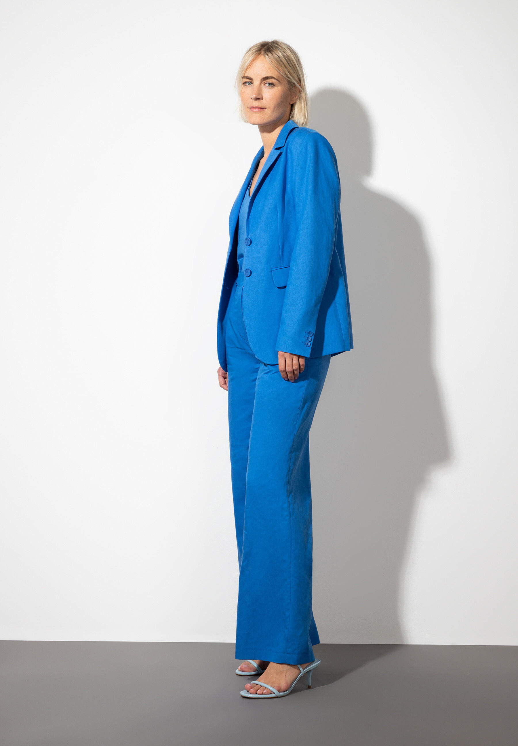 Eine Frau in einem blauen Hosenanzug von MORE & MORE steht vor einer weißen Wand. Sie trägt dazu passende Schuhe mit Absatz. Eine Frau in einem blauen Hosenanzug von MORE & MORE steht vor einer weißen Wand. Sie trägt dazu passende Schuhe mit Absatz.