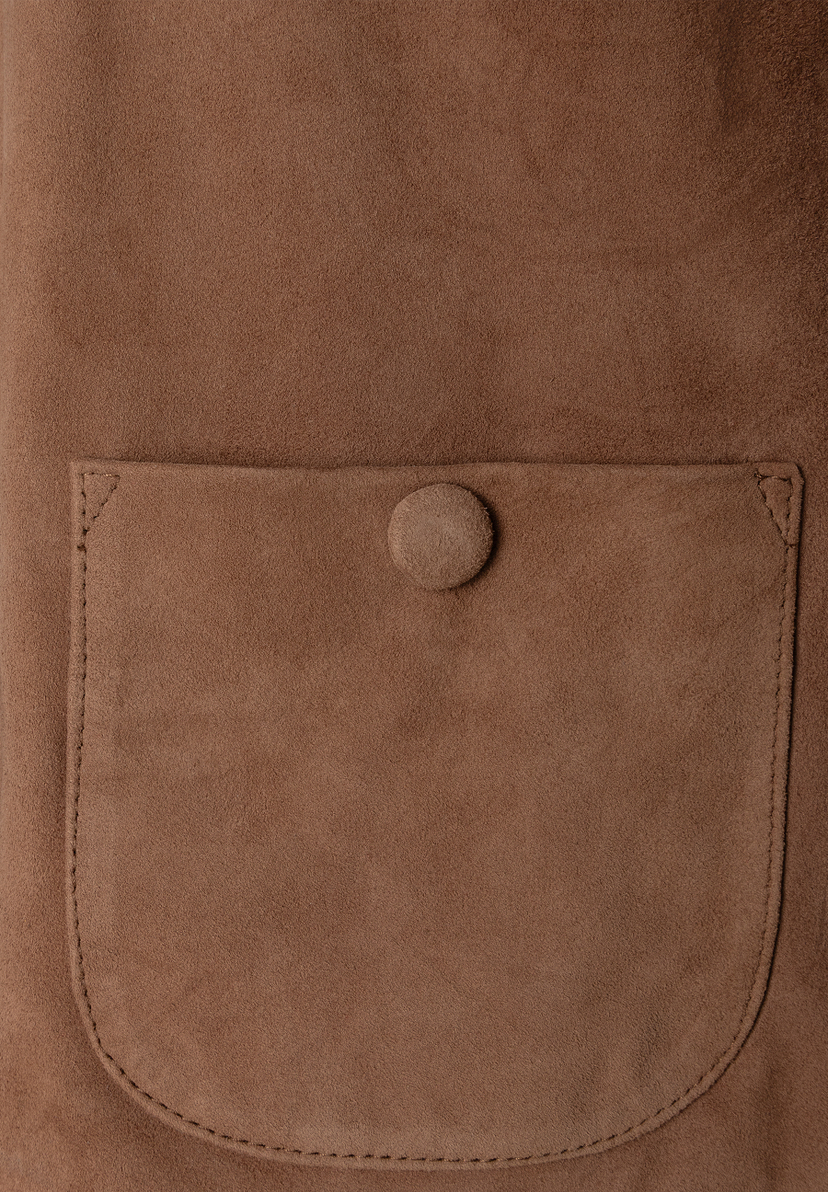 Nahaufnahme eines MORE & MORE Oberteils aus braunem Veloursleder mit aufgesetzter Tasche und Knopf. Nahaufnahme eines MORE & MORE Oberteils aus braunem Veloursleder mit aufgesetzter Tasche und Knopf.